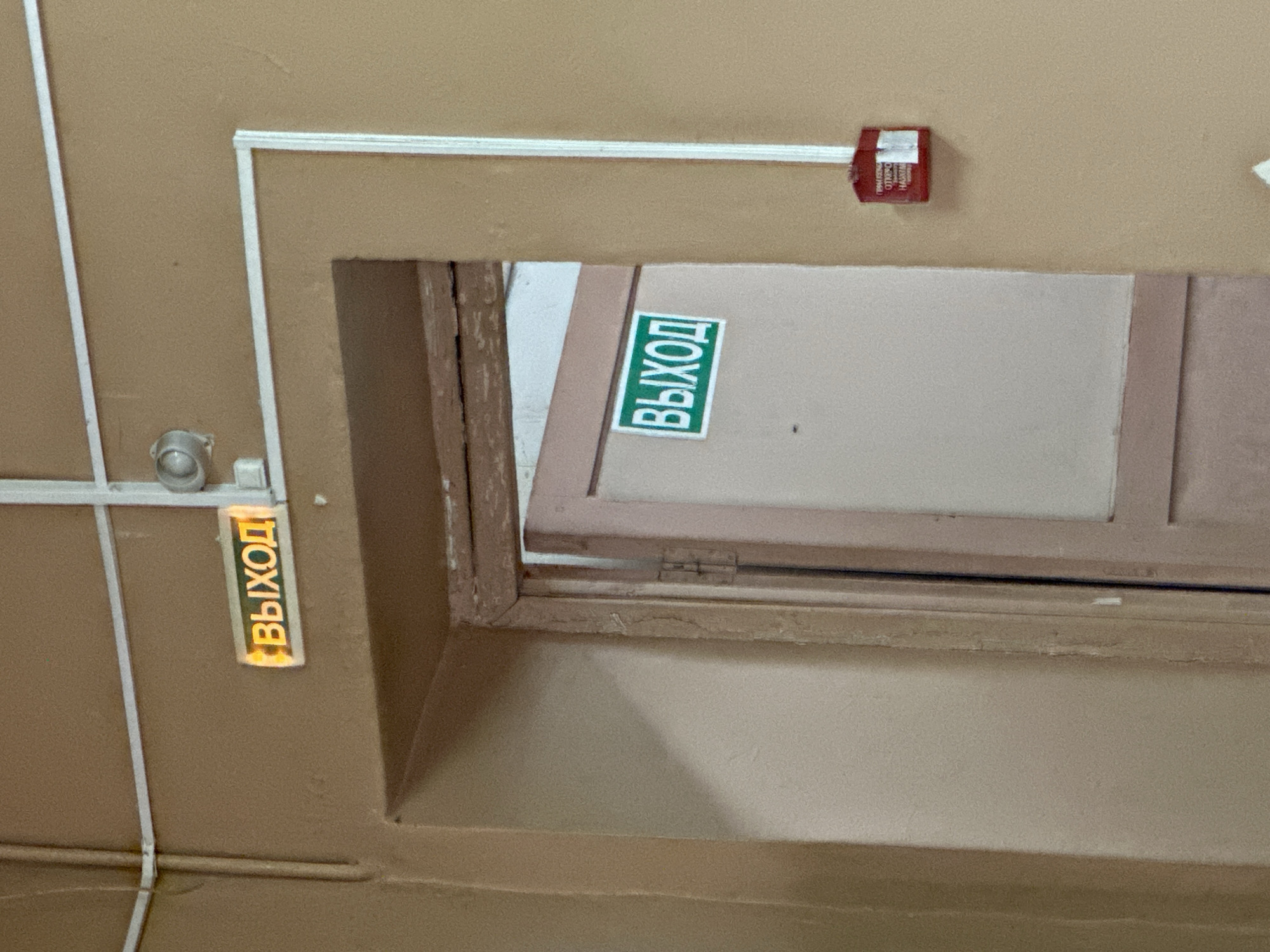 The image shows a hallway with two illuminated exit signs. One sign, located above a doorway, displays the word "ВЫХОД" (Exit) in yellow lettering on an orange background. Below this sign, the doorway is partially open, revealing a door with another "ВЫХОД" sign in green lettering on a white background. To the right of the doorway, a red fire alarm pull station is mounted on the tan-colored wall. The hallway appears to be inside a building, potentially a public space or institution. The lighting is dim, and the walls are painted a neutral color. The overall impression is one of an ordinary interior corridor. The image was taken in Gavrilov Posad, Russia, suggesting the context of a building within that city.