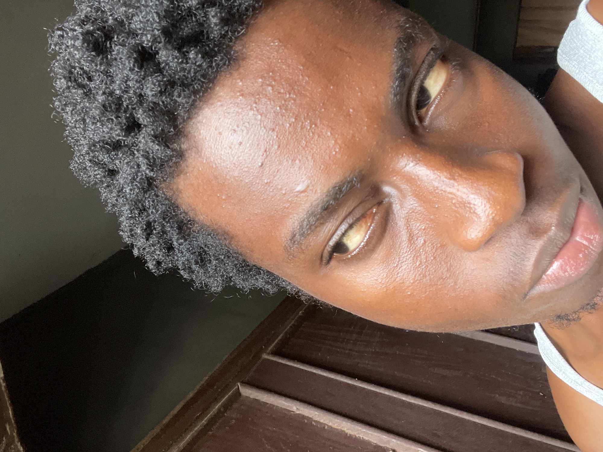 This media file features a close-up, angled shot of a person's face, with the camera looking down on them. The person has dark skin and short, curly black hair. Their face appears slightly sweaty, with visible pores and a sheen on the skin, suggesting warm or humid conditions. The lighting indicates it is daytime, possibly indoors or in a shaded outdoor area, as there are no harsh shadows.
The background is out of focus, but shows a dark, indeterminate space on the left and a wooden surface with horizontal planks on the right. The wooden planks appear to be part of furniture or a structure, possibly a deck or a bench. The person is wearing a white strap, likely from a tank top or undershirt, visible on the upper right side of the frame.
The person's expression is neutral, with their eyes open and looking away from the camera. The overall mood is candid and personal. The location context provided places this scene in Nkpor, Nigeria, which aligns with the visual cues of potentially warm and humid weather. No text is visible in the image.