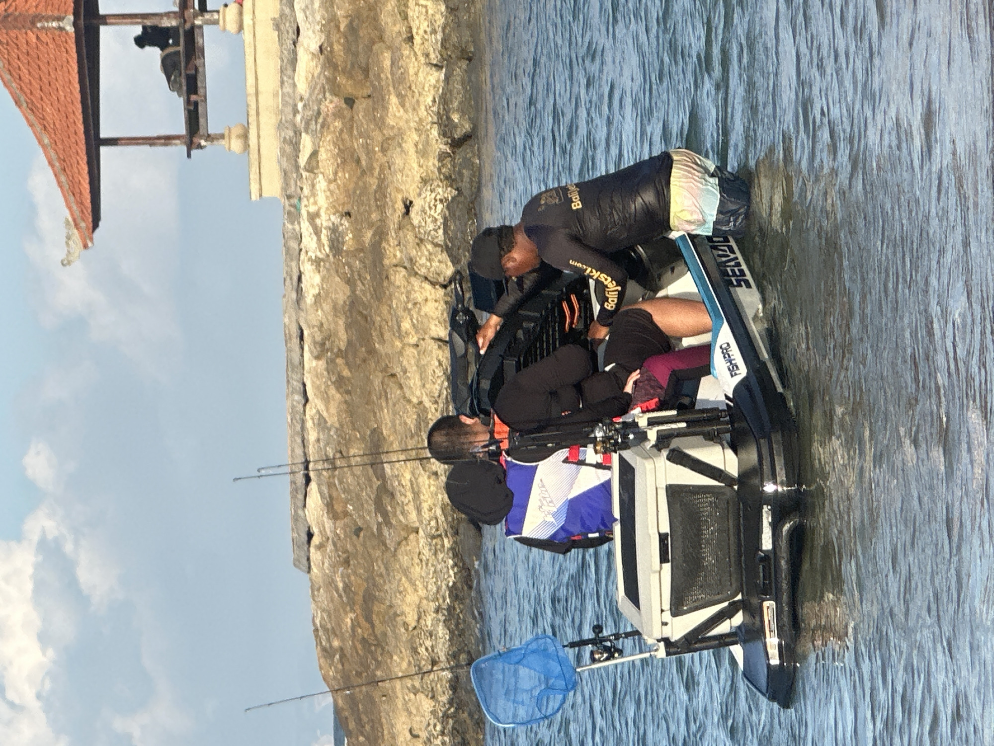 Three individuals are on a jet ski in the ocean near a rock jetty and a covered structure. One person, wearing a black long-sleeved shirt and a baseball cap, stands in the water and appears to be operating or assisting with the jet ski. Another person, seated on the jet ski, is wearing a blue and white life vest. A third person, also seated, is wearing a black long-sleeved top and maroon shorts. Fishing rods and a blue net are attached to the jet ski, which has a cooler mounted on the back. The water is calm and clear. The background features a stone breakwater and a traditional Balinese-style gazebo with a red roof. The location is the waters off the coast of Denpasar, Indonesia.