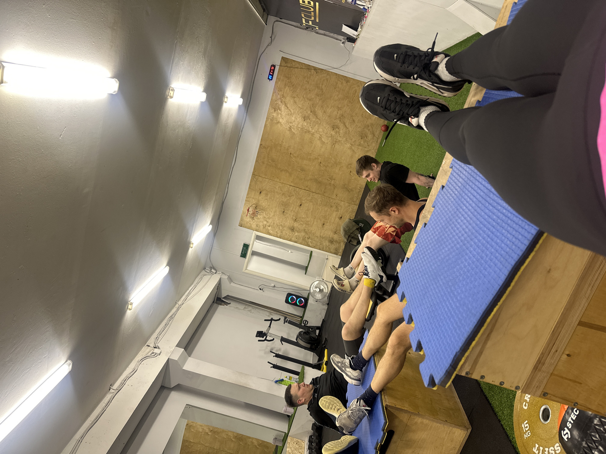 This low-angle photograph captures an indoor fitness studio or gym in Chaikovsky, Russia. The perspective is from someone lying or sitting on a blue exercise mat atop a wooden plyo box, with their legs and feet (wearing black leggings and black Nike sneakers) prominently in the foreground.
Four individuals, mostly men, are visible lying on their backs with their legs elevated on similar wooden boxes topped with blue interlocking foam mats. They appear to be either resting or performing an exercise such as core work or stretching. One man in the mid-ground wears a black t-shirt and grey sneakers.
The gym features bright fluorescent tube lights along the white walls and ceiling. One wall is clad in light wooden paneling, while another is white with an emergency exit sign. The flooring alternates between green artificial turf and darker matting. Visible gym equipment includes a rowing machine, a standing fan, and a red medicine ball. A digital clock or timer displays "31:18" on the wooden wall. A partial black and gold sign with the text "FCLUB" is also visible. On the floor, a 15 kg weight plate with "STEC" branding can be seen. The scene depicts a functional fitness environment.