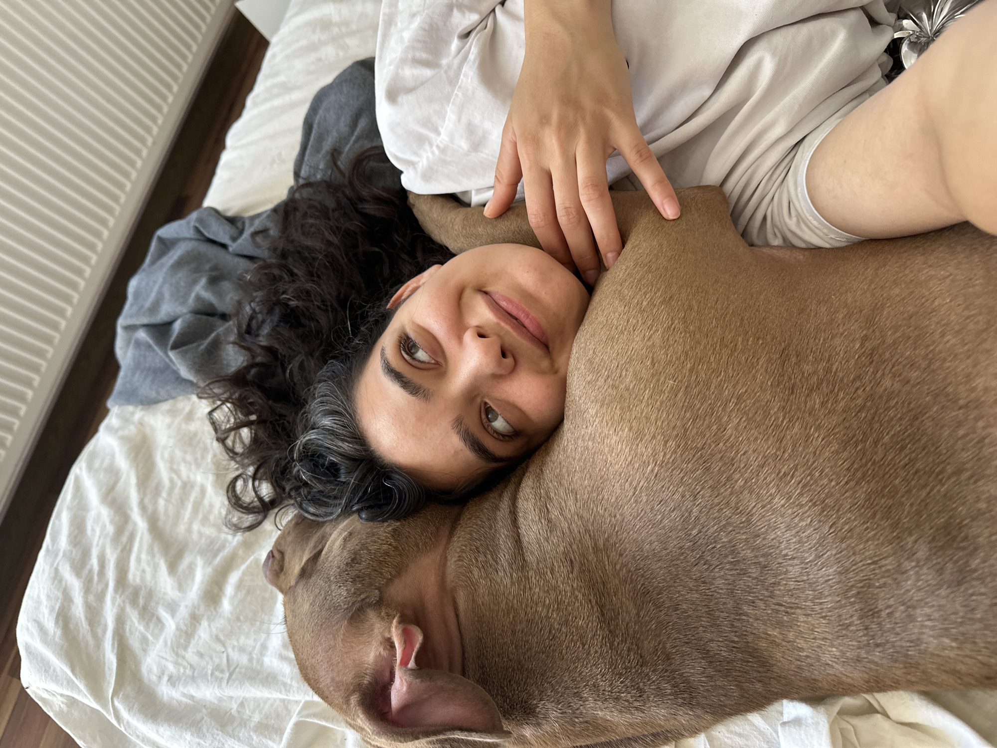 A young woman with dark, curly hair is lying in bed, embracing a light brown dog. The woman's head is tilted towards the dog, and she is smiling. Her right arm is wrapped around the dog's neck, and her hand rests on its shoulder. The dog is lying on its side, with its head resting on the woman's chest. The scene takes place in a bedroom, with white sheets on the bed and a grey blanket in the background. The lighting is soft and natural. The image appears to have been taken from a high angle, looking down on the woman and dog. The location is identified as Malevo, Bulgaria, though no specific city landmarks are visible.