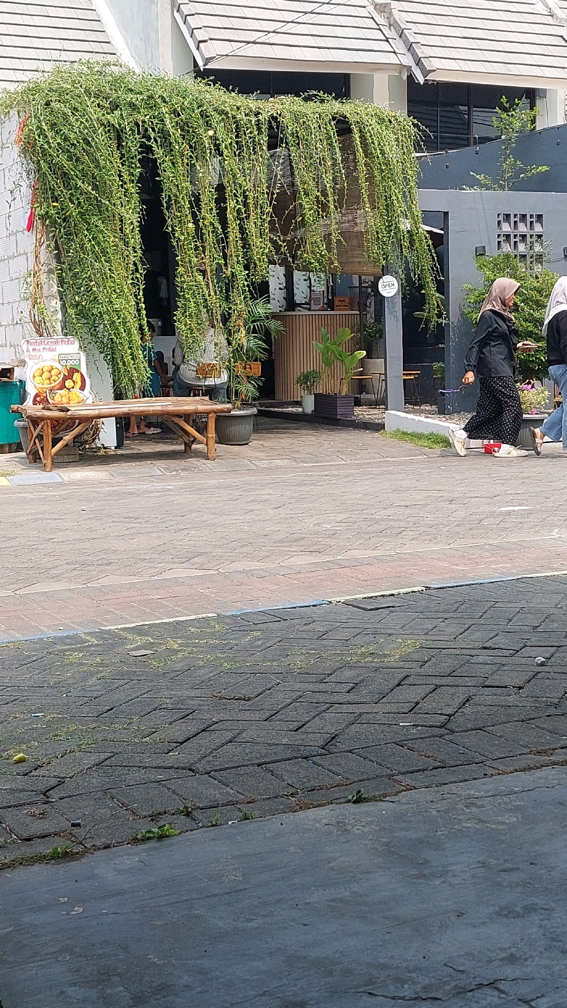 The image captures a street-level view of a building facade adorned with lush, hanging green foliage. A woman wearing a black hijab and a patterned dress, accompanied by another person in the background, is walking on a paved walkway in front of the building. To the left, a bamboo bench sits in front of a display board featuring images of food items and text in Indonesian, suggesting a commercial establishment. The building itself appears to be a shop or cafe, with its entrance partially obscured by the greenery. The ground is paved with interlocking bricks, transitioning to a smoother concrete surface in the foreground. The overall scene suggests a vibrant streetscape in a residential or commercial area, likely in a tropical climate given the abundant plant life. The presence of the Indonesian text on the display board indicates the location is in Indonesia, and specifically, the text mentions "Jimbaran Kulon," placing the scene in that area.