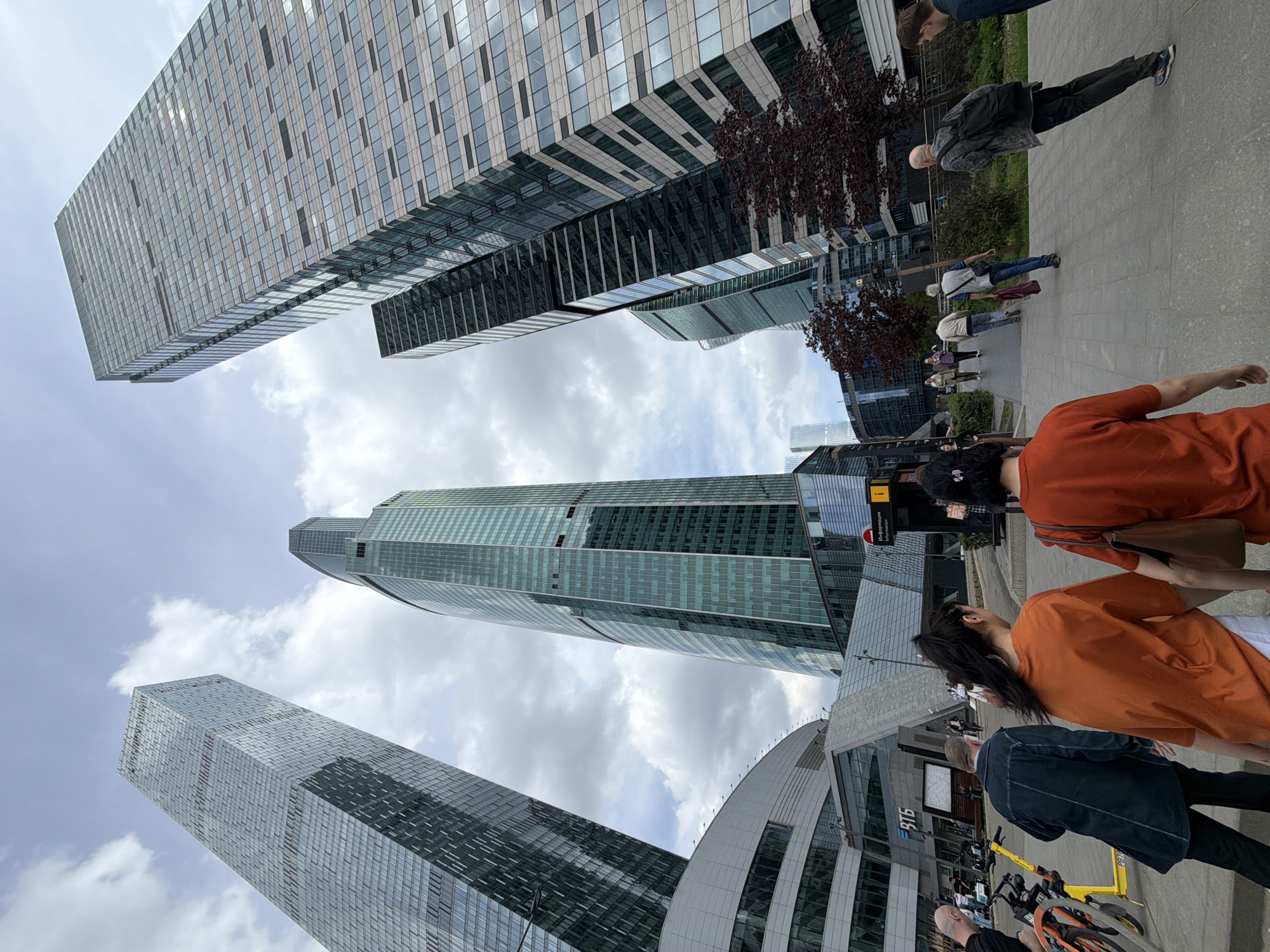 The image captures a street scene in Khimki, Russia, featuring several modern skyscrapers. The buildings are predominantly glass and steel, reflecting the sky and clouds. Pedestrians walk along a paved area adjacent to the buildings. Some are wearing casual clothing. There are parked bicycles visible. A small tree with dark leaves is situated near the buildings. The sky is partly cloudy.