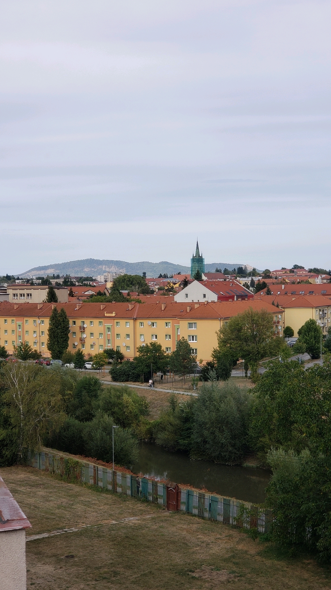 This is an elevated, full-frame shot of a residential area in Prešov, Slovakia, on an overcast day. In the foreground, a grassy area with a patterned path leads to a weathered metal fence bordering a dark, slow-moving body of water, possibly a canal or a narrow river. The fence is a patchwork of green, brown, and red panels, with some sections appearing rusted. Beyond the water, there are lush green trees and bushes lining the embankment.
Further back, several apartment buildings with orange tiled roofs dominate the midground. The buildings are multi-story, painted in various shades of yellow and cream, and feature numerous windows, some with satellite dishes attached. A street runs in front of these buildings, with a few parked cars visible. Two individuals are walking on the sidewalk near the street.
In the background, a cluster of buildings with red roofs is visible, interspersed with trees. A prominent church spire, partially covered by green scaffolding, rises above the rooftops. In the far distance, rolling hills and mountains form the horizon under a pale, hazy sky. The overall impression is of a calm, possibly suburban, setting within the city.