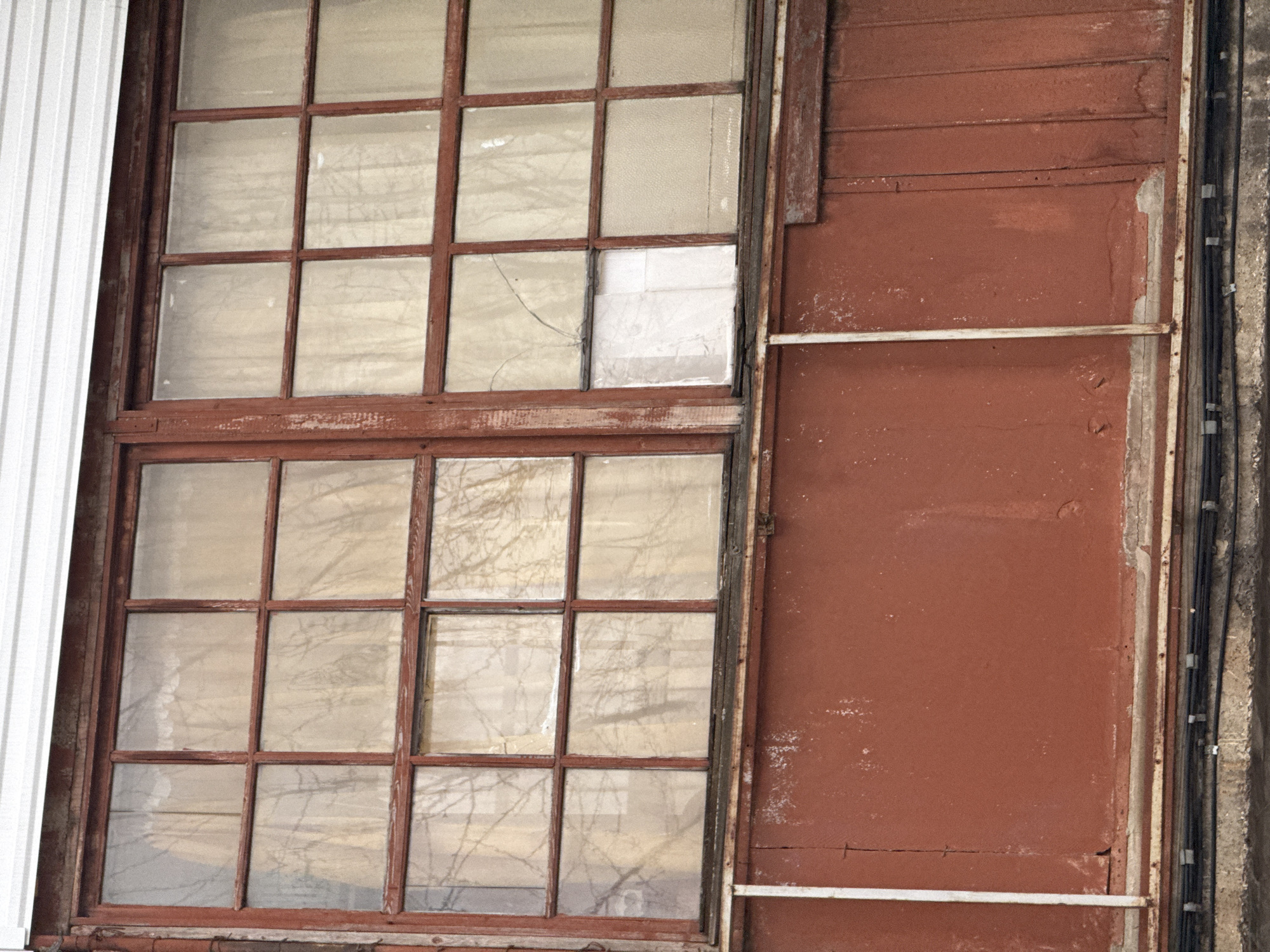 The image shows a weathered building exterior, likely in Perm, Russia, given the request. A large, divided-pane window dominates the left side of the frame, with a red-brown wooden frame. One pane is cracked. The window reflects indistinct shapes, possibly trees. To the right is a rusted red-brown wall with horizontal boards, marred by peeling paint. A white metal beam is mounted to the side. On the far right are several black electrical cables running vertically along the wall, encased in conduit. The building appears old and unkempt. On the far left is white corrugated metal siding.
