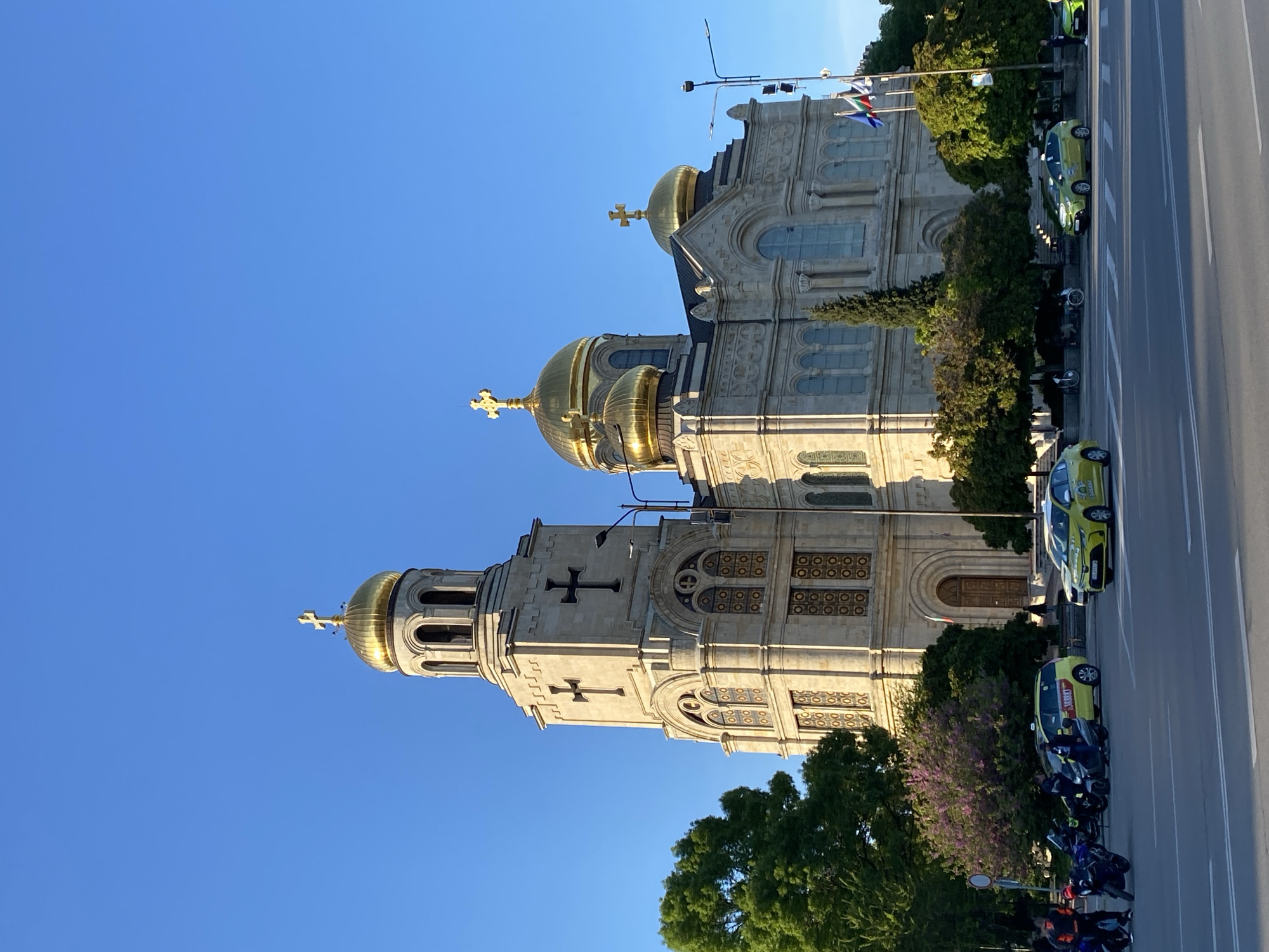 The image shows the Cathedral of the Assumption of the Virgin in Varna, Bulgaria, from a ground-level perspective. The architectural details of the cathedral, with its golden domes and crosses, are visible against a clear blue sky. Trees partially obscure the lower portion of the building. A road runs alongside the cathedral, with several parked yellow and green taxis and motorcycles. A few pedestrians are visible. Flags of the city of Varna are at the top. The time of day appears to be morning or late afternoon, based on the lighting.