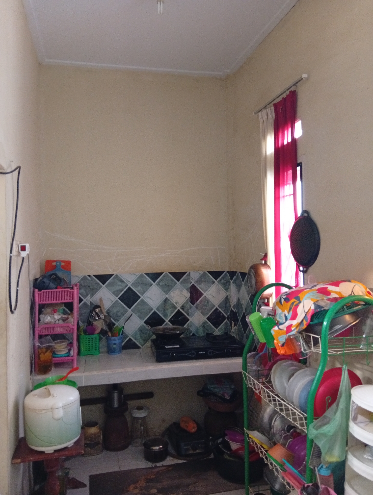 This is an indoor, medium-angle, eye-level shot of a modest kitchen area in Kualu, Indonesia. The walls are painted a neutral, light color, with some chalk-like marks visible. A white ceiling with a visible light fixture is at the top. On the left side, a black cable runs from a white electrical outlet mounted on the wall.
The kitchen counter is tiled with a geometric pattern of green, black, and white squares. On the counter, a pink, multi-tiered shelf unit holds various items, including plastic containers and what appears to be bags of groceries. To the right of the shelf, a small green basket contains utensils, and next to it is a blue basket with more utensils. A gas stovetop is positioned on the counter to the right, with a dark pot or pan on it.
A white rice cooker is situated on a low wooden shelf on the left side of the image, below the counter. Next to it are several dark-colored containers.
In the background, a window is covered by a curtain that is predominantly bright pink, with some white fabric visible. Sunlight streams through the window, illuminating the area. To the right of the window, a dark, circular object, possibly a fan or a griddle, is attached to a stand.
A green dish rack is prominently featured on the right side of the image, filled with various plates, bowls, and cups. A colorful patterned cloth is draped over the top rack of the dish rack. Several plastic bottles, including a green one and a red squeeze bottle, are also present in or around the dish rack. In the lower right corner, a stack of white containers, likely food storage containers, is visible.
The overall impression is of a functional, lived-in kitchen.