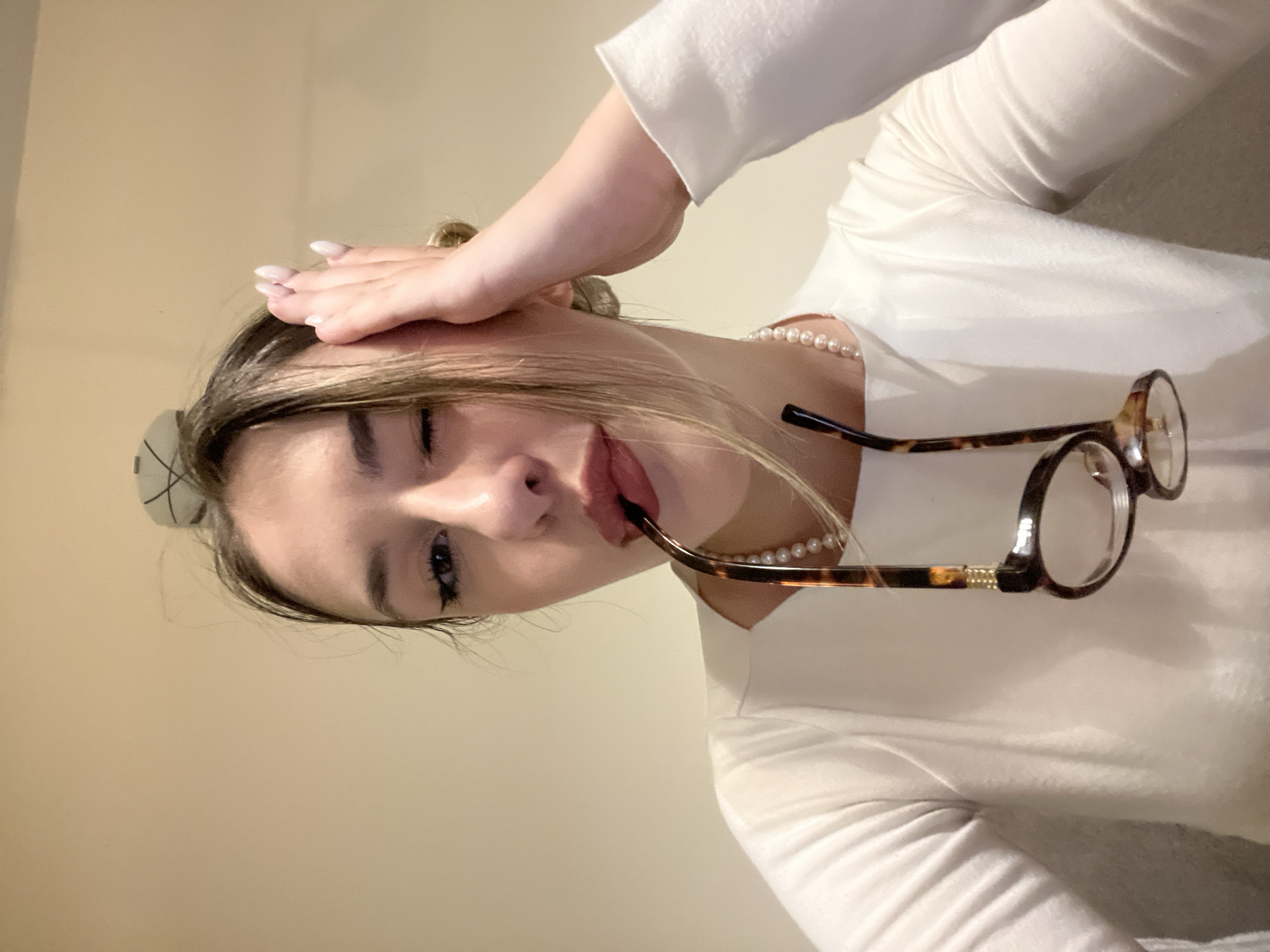 The image captures a young woman from an overhead perspective, with her head tilted sharply to the right. She has light-colored hair, some strands falling across her forehead, and a light-colored hair tie visible at the back of her head. Her right hand, adorned with light-polished, manicured fingernails, rests gently on her forehead.
She is winking with her right eye, and her lips are slightly parted, holding a pair of tortoiseshell-framed eyeglasses by one earpiece. Around her neck, she wears a pearl necklace, partially visible. She is dressed in a white long-sleeved top or shirt. The background consists of a plain, light beige or off-white wall, indicating an indoor setting. The lighting is soft and even. No text is visible in the image.