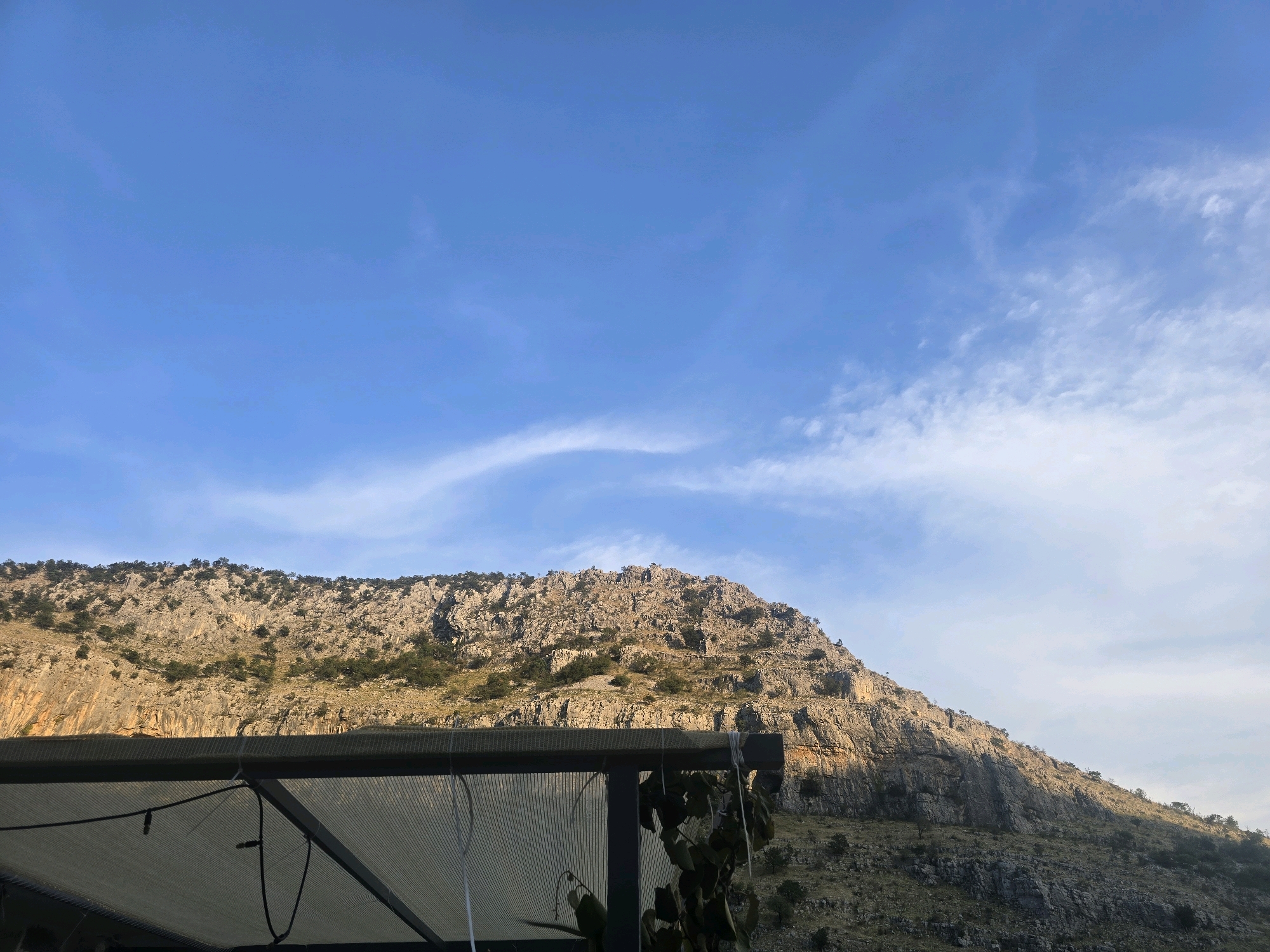 The image shows a mountainous landscape under a blue sky with scattered clouds. In the foreground, a dark metal structure with a light-colored, semi-transparent mesh roof is visible, casting shadows. Some greenery hangs from the structure on the right. The mountain itself is rocky and steep, with sparse vegetation scattered across its slopes. The sunlight illuminates the mountain from the side, creating distinct shadows and highlighting the rugged texture of the rock face. The overall impression is of a natural, rugged environment, possibly near a built structure offering shade. This scene is characteristic of the outskirts or natural areas surrounding Podgorica, Montenegro, where mountainous terrain is readily accessible.