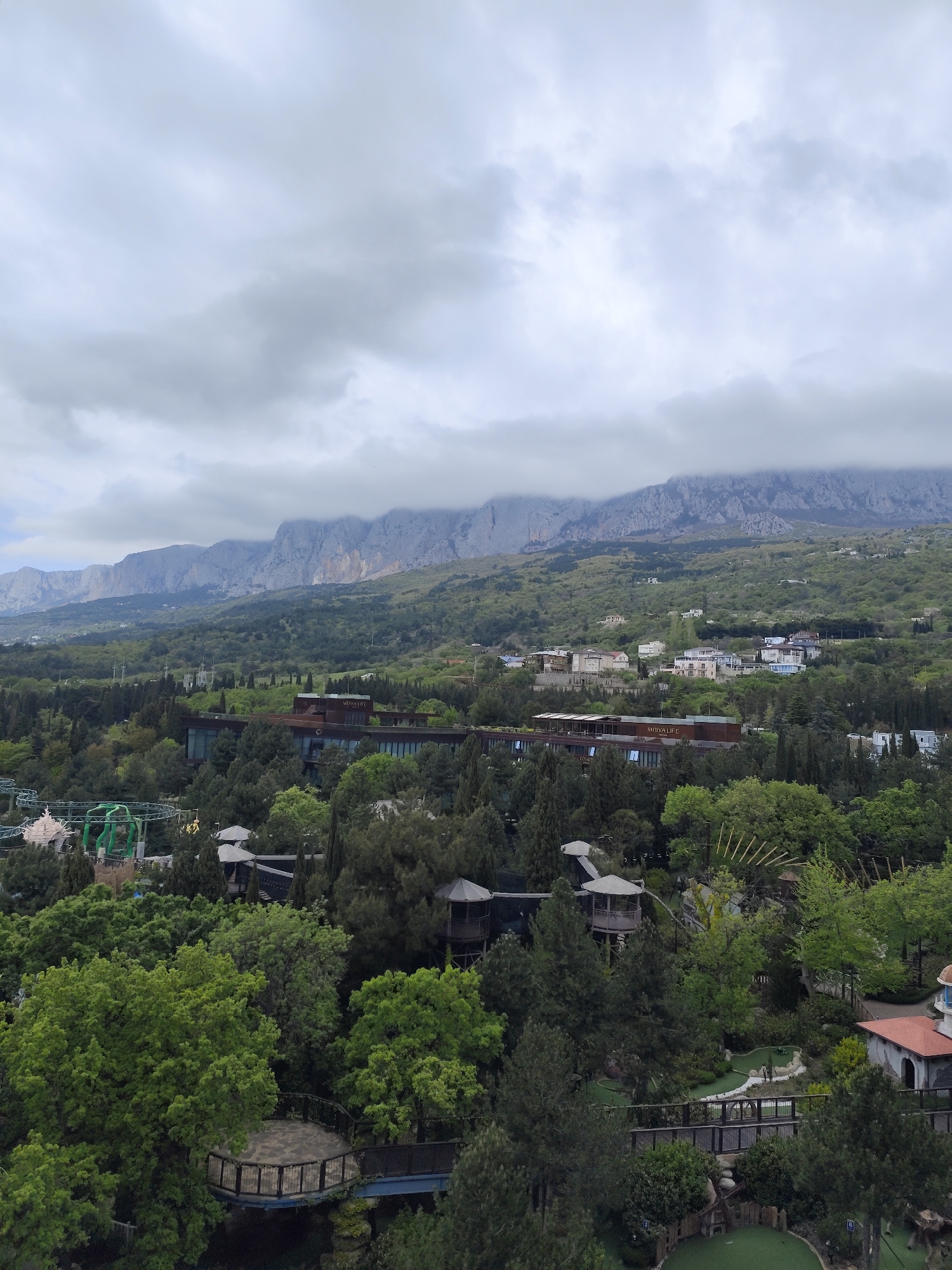 The image shows a landscape in Симеизский поселковый совет, Ukraine. Mountains are visible in the distance, partially obscured by low-hanging clouds. Below the mountains, there is a valley filled with dense green trees. A large modern building is nestled within the trees. In the foreground, there are structures resembling treehouses and a miniature golf course. A roller coaster is visible on the left.