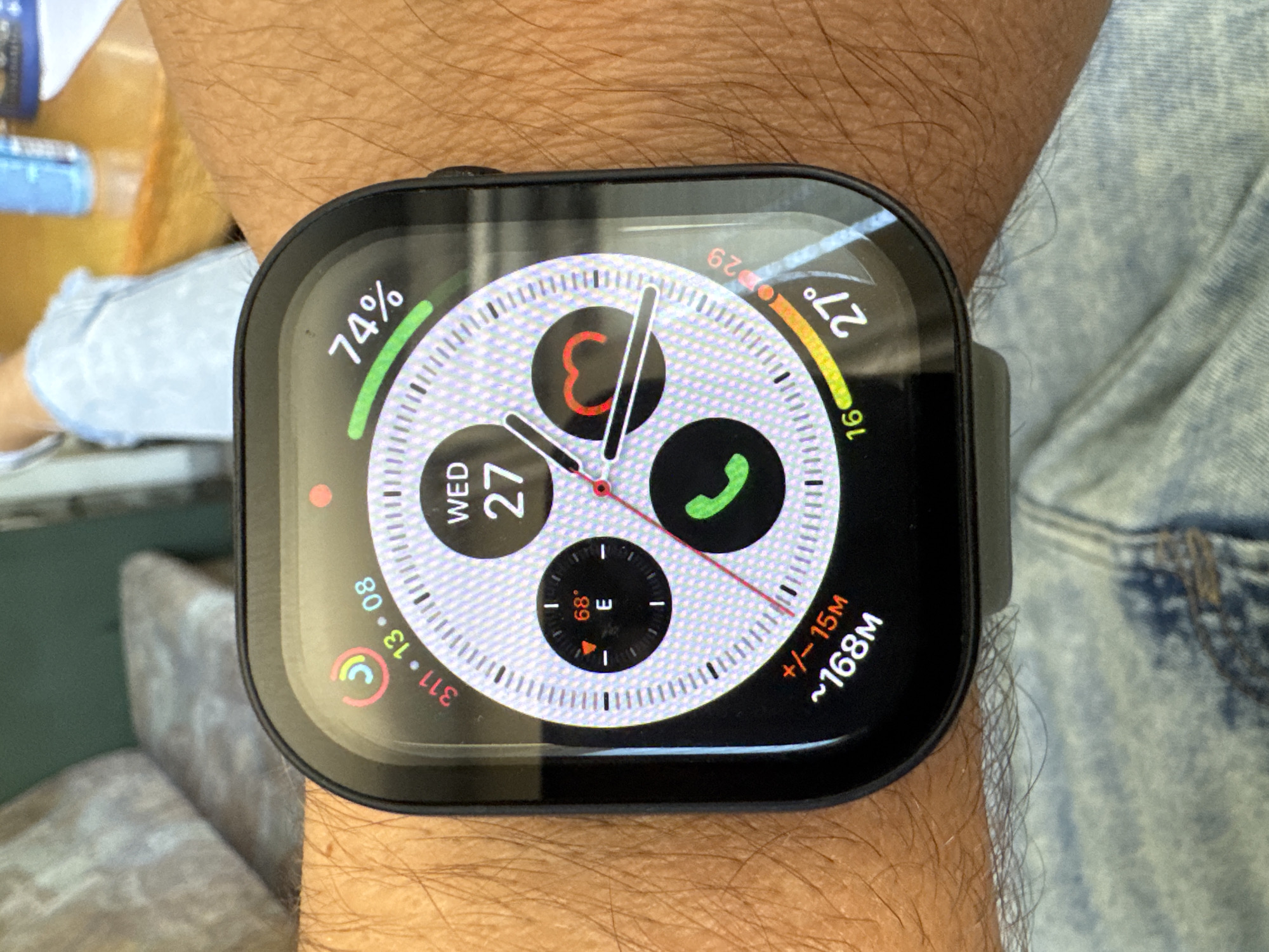The image shows a person's wrist with an Apple Watch. The watch face displays the time, date (Wednesday the 27th), weather (27°C), battery level (74%), compass direction (East), and some other icons representing phone calls and messages. In addition, the display indicates a measurement of approximately 168M with a variance of +/-15M. The surrounding environment shows the person's arm, clothing (jeans), and glimpses of furniture in the background. It is not possible to confirm the exact location (Plovdiv, Bulgaria) based solely on the visual information provided.