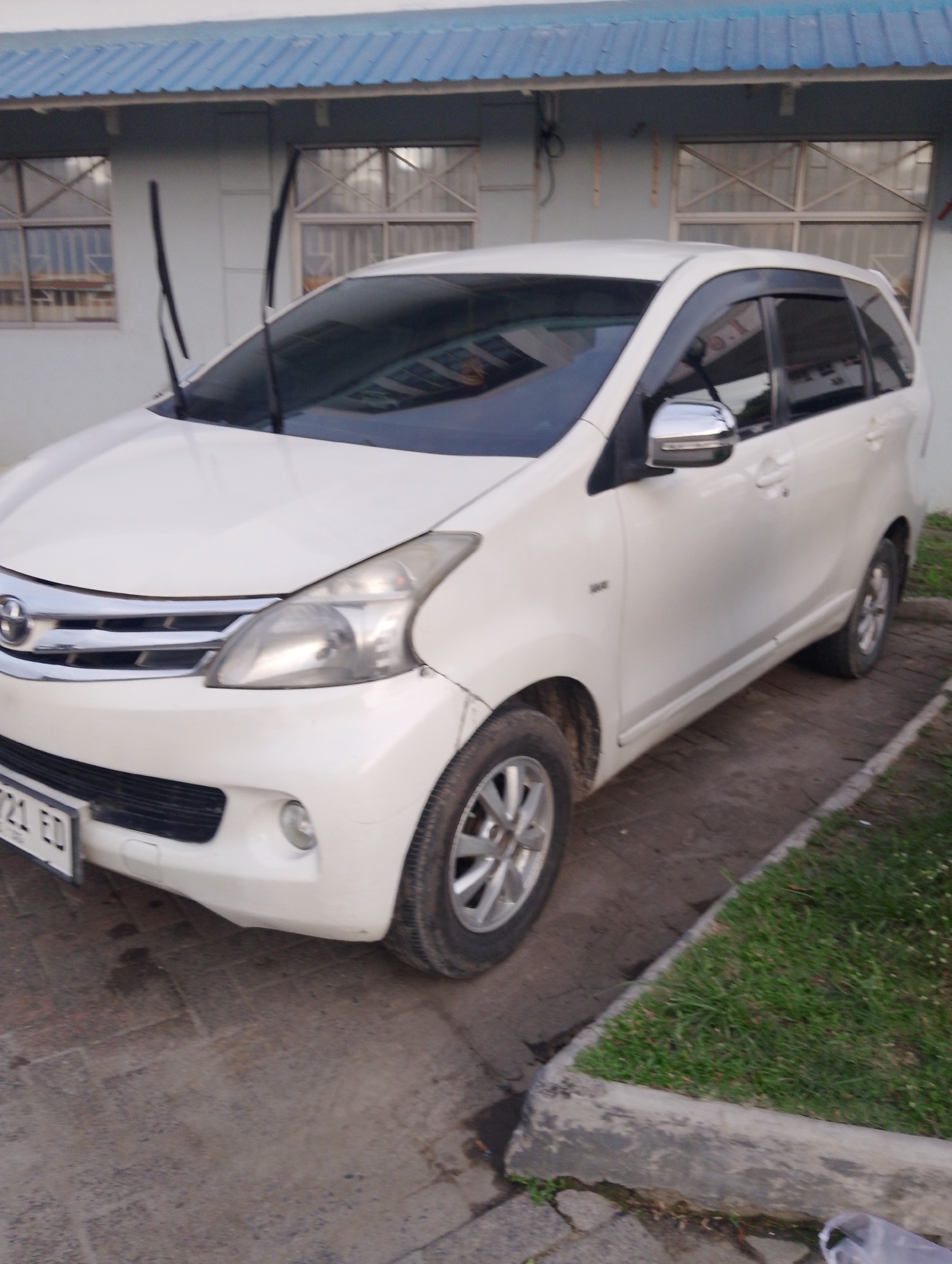 A white Toyota Avanza is parked in front of a building with a blue corrugated metal roof and two windows with metal frames and blinds. The car has a license plate that reads "B 721 ED". There are two windshield wipers visible on the car. The car is parked on a paved surface next to a patch of grass. The environment appears to be an urban setting, potentially in the City of Pematang Siantar, Indonesia, based on the license plate format.