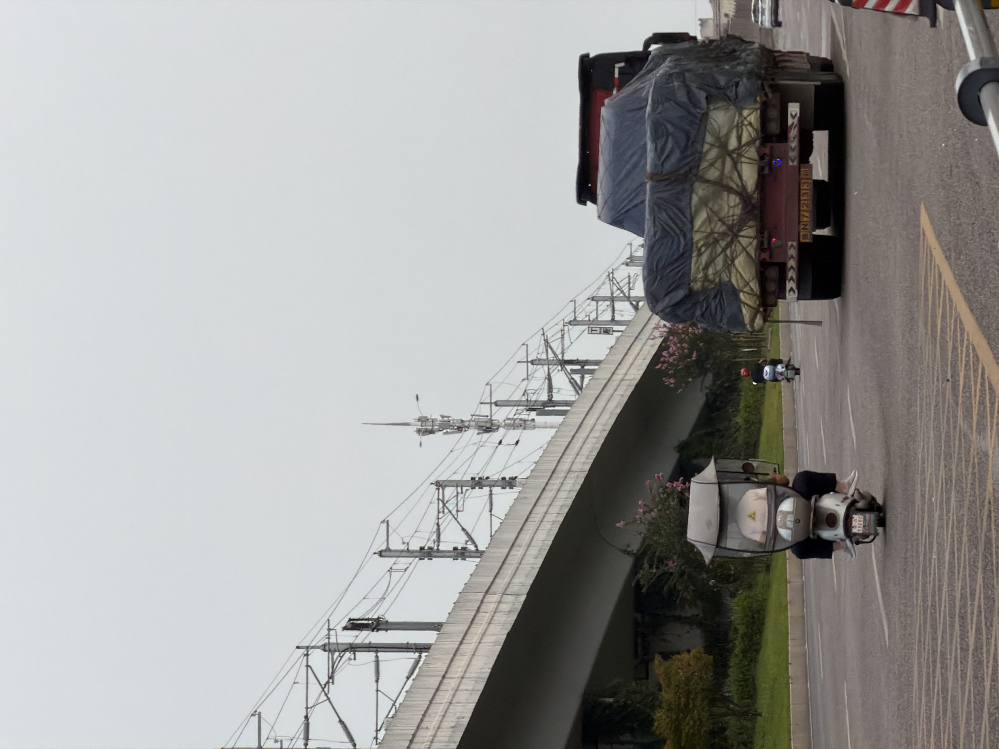 The image depicts a street scene in Jiangjin, China, under an overcast sky. In the foreground, a person on a scooter with a canopy is traveling on the road. The scooter has a visible license plate with Chinese characters and numbers. Further down the road, a large truck is visible, its cargo covered by a dark tarp secured with ropes. The truck also has a visible license plate. In the background, an elevated structure, possibly a highway or train track, is supported by concrete pillars and crisscrossed with overhead electrical wires. Greenery and trees are present on the left side of the road. The overall atmosphere is one of ordinary daily life in an urban environment.