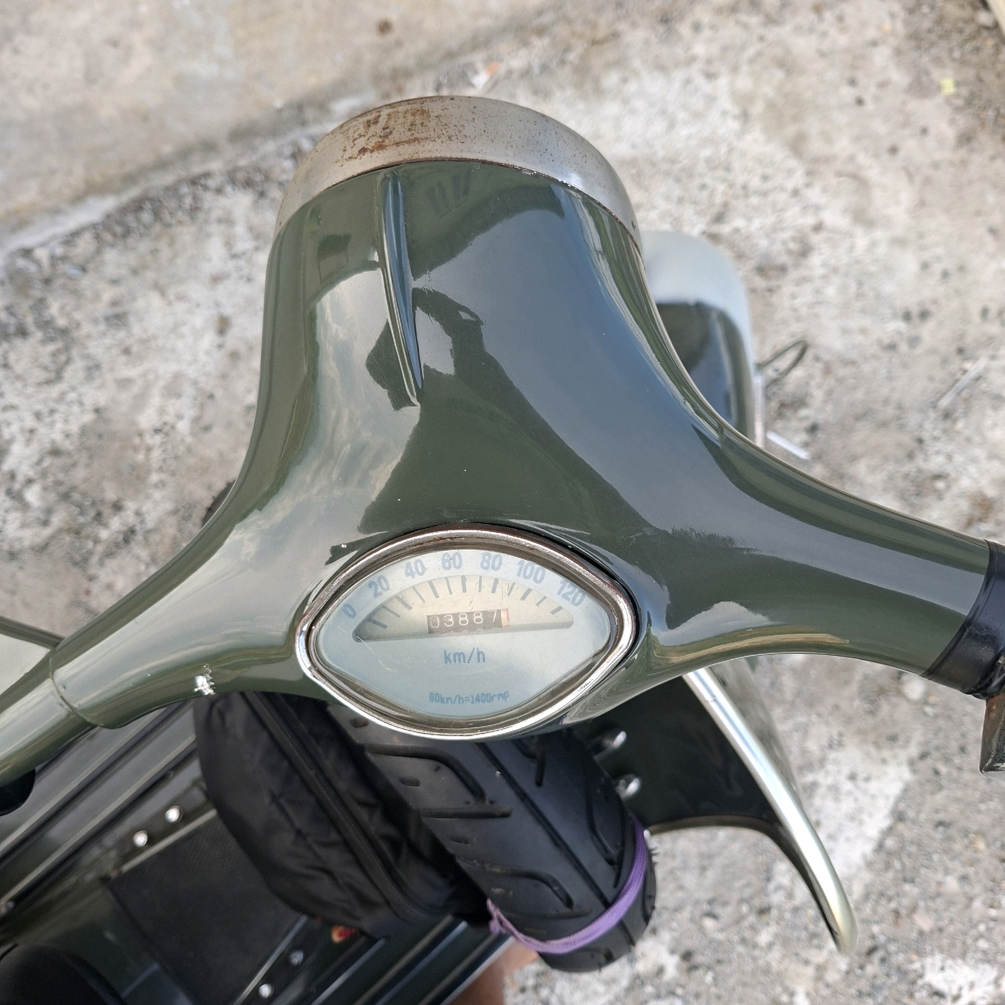 The image shows a close-up, high-angle view of a vintage green scooter's handlebars and instrument cluster, set against a textured, light-colored concrete surface, possibly a street or sidewalk. The scooter's handlebar is glossy and reflects the sky and surrounding environment. The instrument cluster features a speedometer with markings up to 120 km/h, displaying a mileage of "03887". Below the speedometer, the text "km/h" is visible, along with "60km/h=1400rpm" on a smaller dial. A black bag is attached to the side of the scooter, secured with a purple tie-wrap around the front wheel. The foreground shows the front tire of the scooter. The background consists of a rough concrete surface with scattered pebbles and debris. The location is identified as Gronggrong, Indonesia. There are no people visible in the image.