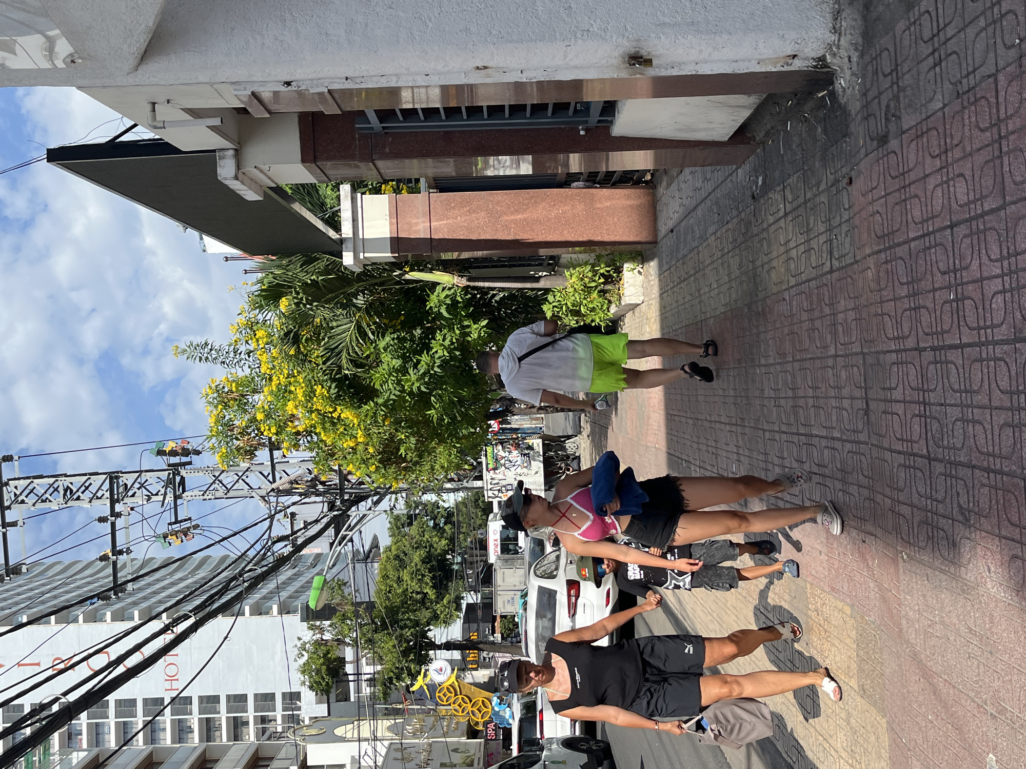 A wide shot captures a bustling street scene in Nha Trang, Vietnam, on a bright, sunny day. A four-lane road, lined with parked and moving vehicles, runs parallel to a wide, tiled sidewalk. The sidewalk is populated by several individuals, including a woman in a black tank top and black shorts, holding a beige tote bag, and a child in a grey and white graphic t-shirt and grey shorts. They appear to be walking together, possibly as a family. To their right, a young woman in a pink and white crochet halter top and black shorts walks alongside them, carrying a blue towel. Further ahead, a man in a white t-shirt and bright lime green shorts walks away from the camera, his back to the viewer.
The environment is urban, characterized by tall buildings, including a prominent modern hotel with numerous balconies, and an extensive network of electrical wires crisscrossing the sky. Lush green trees with vibrant yellow flowers are visible, adding a splash of color to the scene. The architecture includes decorative stone pillars and a large, dark electronic billboard mounted high on a building. The sidewalk itself is paved with red and beige square tiles in a patterned arrangement. Debris, including what appears to be cigarette butts, is scattered along the edge of the sidewalk near a building entrance. The lighting suggests it is midday, with strong shadows cast by the sun.