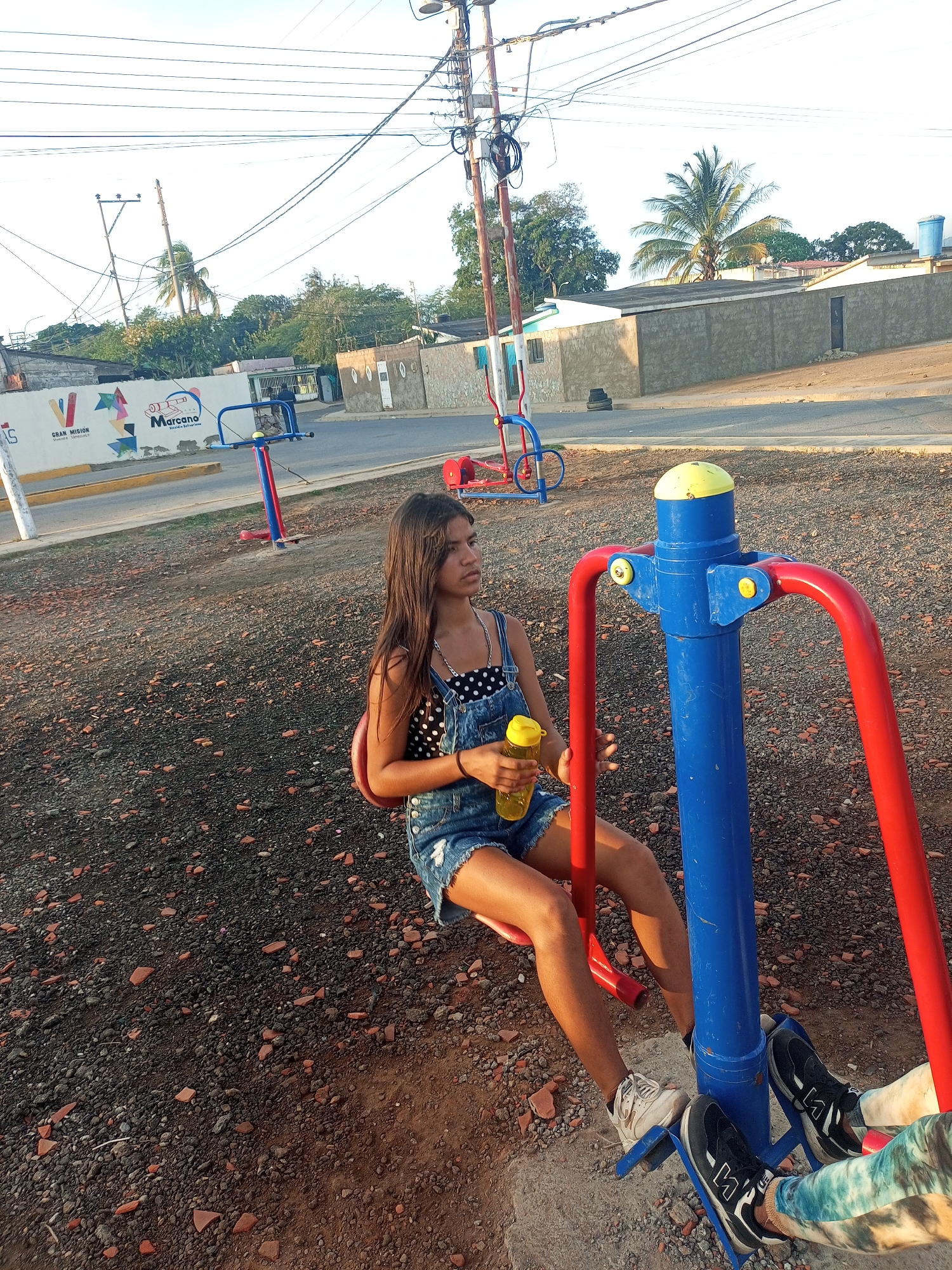 A young woman with long brown hair and wearing a polka dot top and denim overalls is seated on a blue and red outdoor exercise machine, holding a yellow water bottle. The machine is located in an outdoor space covered in dark gravel and small reddish stones. Behind her, other similar exercise machines can be seen, along with a painted wall displaying the words "GRAN MISIÓN" and "Marcano". The location is Juan Griego, Venezuela. The background also shows houses, trees, and a palm tree, under a bright sky. Another person’s legs and feet can be seen on the exercise machine as well, suggesting that the subject is interacting with them on the machine.