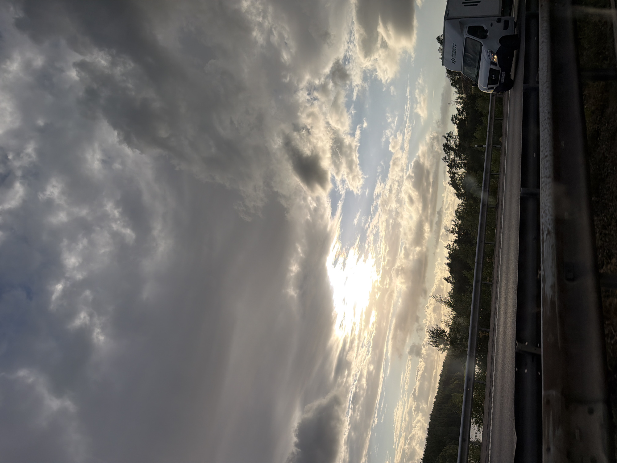 The image captures a roadside view near Kläppa, Sweden, on a cloudy day with the sun breaking through the clouds. A white cargo van with " Rydbergs Trävaror" signage on its side is traveling on a highway. The van is on the right side of the frame, moving away from the viewer. The highway is bordered by a metal guardrail in the foreground and a more substantial guardrail separating the road from a wooded area. The background features a dense forest line along a hill, with trees and vegetation. The sky is filled with dramatic, heavy clouds, but a bright patch of sunlight is visible in the upper left quadrant, illuminating the scene with a warm glow. The overall impression is a snapshot of a journey through a natural landscape under a dynamic sky.
