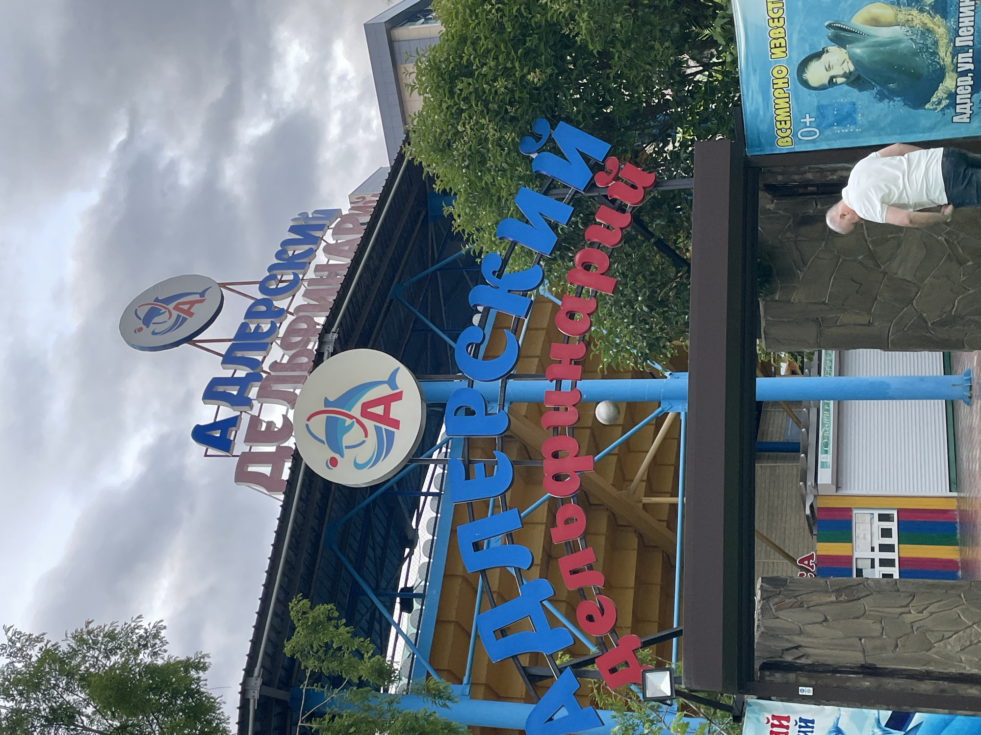 This is a close-up, eye-level, outdoor medium shot of the entrance to a dolphinarium in Sochi, Russia, under a cloudy sky. Dominating the frame is a large sign with large, multi-colored block letters. The top line of the sign, in pink, reads "АДЛЕРСКИЙ" (Adlersky). Below this, the larger blue letters spell out "ДЕЛЬФИНАРИЙ" (Dolphinarium). A circular logo with a dolphin and the letter "A" is positioned above the pink text. In the background, green trees and a modern building with a gray facade are visible. To the right, a blue banner displays a woman swimming with a dolphin and the text "ВСЕМИРНО ИЗВЕСТН" (World Famous) and "0+". Below the banner, street-level signage indicates "Адлер, ул. Ленин". In the foreground, to the left, a stone wall surrounds an entrance area with a ticket booth painted in bright vertical stripes of red, yellow, green, blue, and purple. A man in a white polo shirt and dark pants is walking away from the camera, partially obscuring the entrance. Another man is visible through a window of the ticket booth.