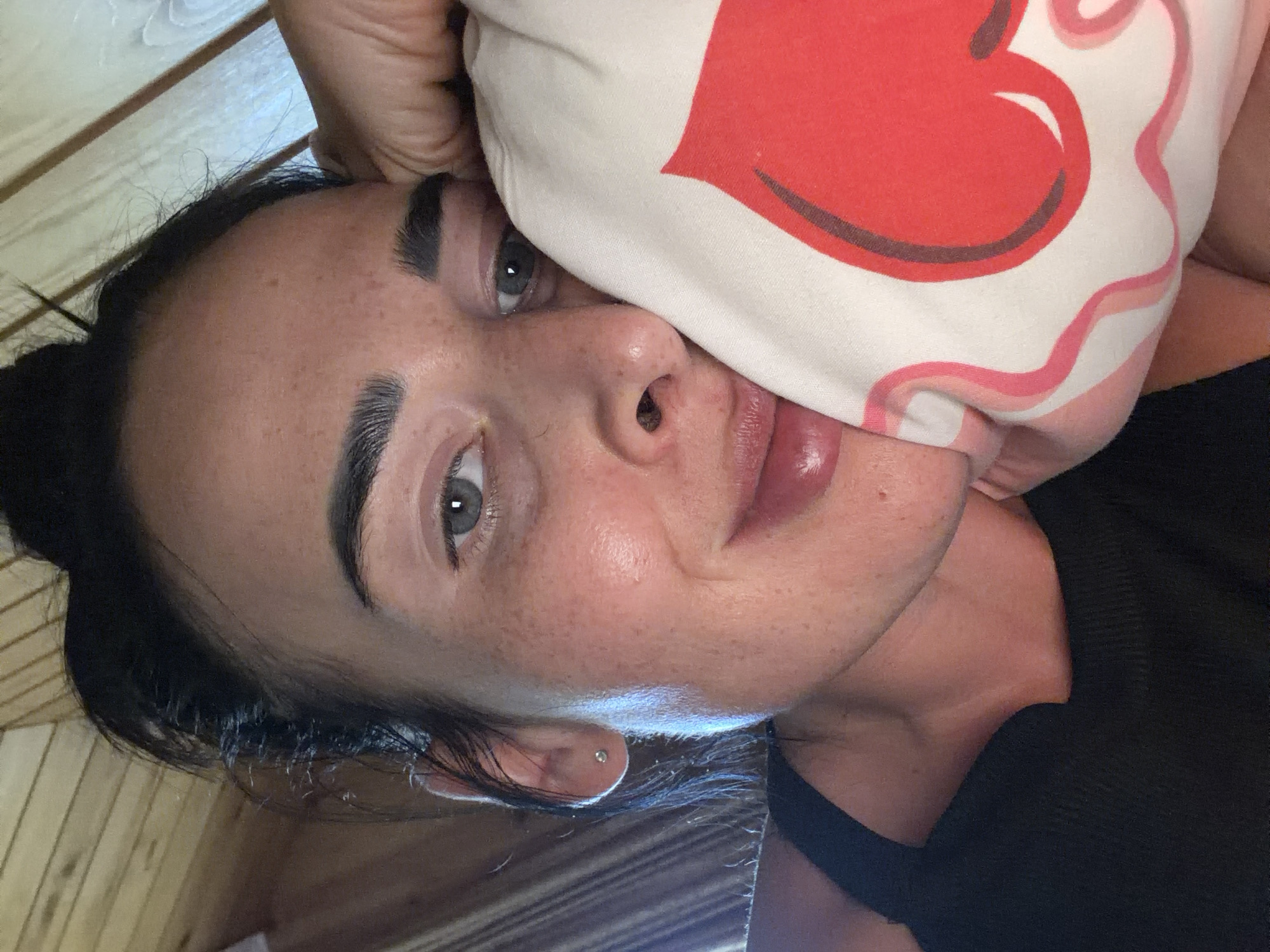 This is a close-up, vertically oriented, tilted photo of a woman's face. She appears to be lying down, with her head propped on a pillow that has a large red heart design. Her left eye is visible, with a light gray iris and dark eyebrows. Her nose is slightly turned away from the camera, and her lips are full and colored with a glossy lipstick. She has a fair complexion with visible freckles across her cheeks and nose. Her dark hair is partially visible, framing her face. A small silver stud earring is visible in her left earlobe. The background appears to be made of wooden planks, suggesting an indoor setting. The lighting is soft, casting a slight sheen on her skin. The woman's expression is relaxed and somewhat alluring. The photo was taken in Sochi, Russia.
