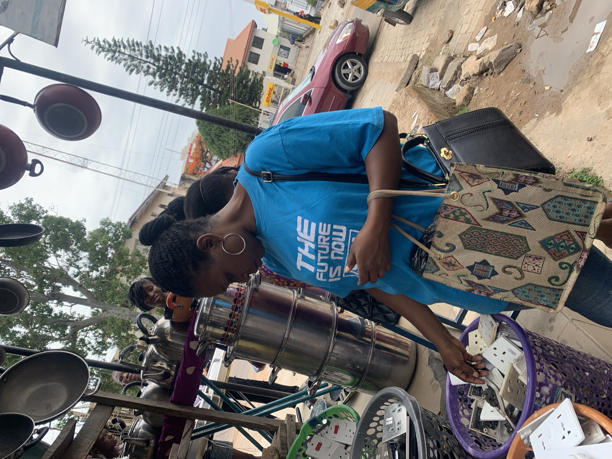 The image depicts an outdoor scene in Jos, Nigeria. A woman wearing a blue t-shirt with the words "THE FUTURE IS NOW" is looking at a purple basket filled with electrical outlets. She is reaching into the basket with her right hand. She is also wearing silver hoop earrings, and carrying two purses. One purse is tapestry-like. There is a rack of cookware behind her, including pots and pans. In the background, there is a red car parked near a building that has a DHL sign. There is street debris and a puddle near the car. Other people can be seen in the background as well.