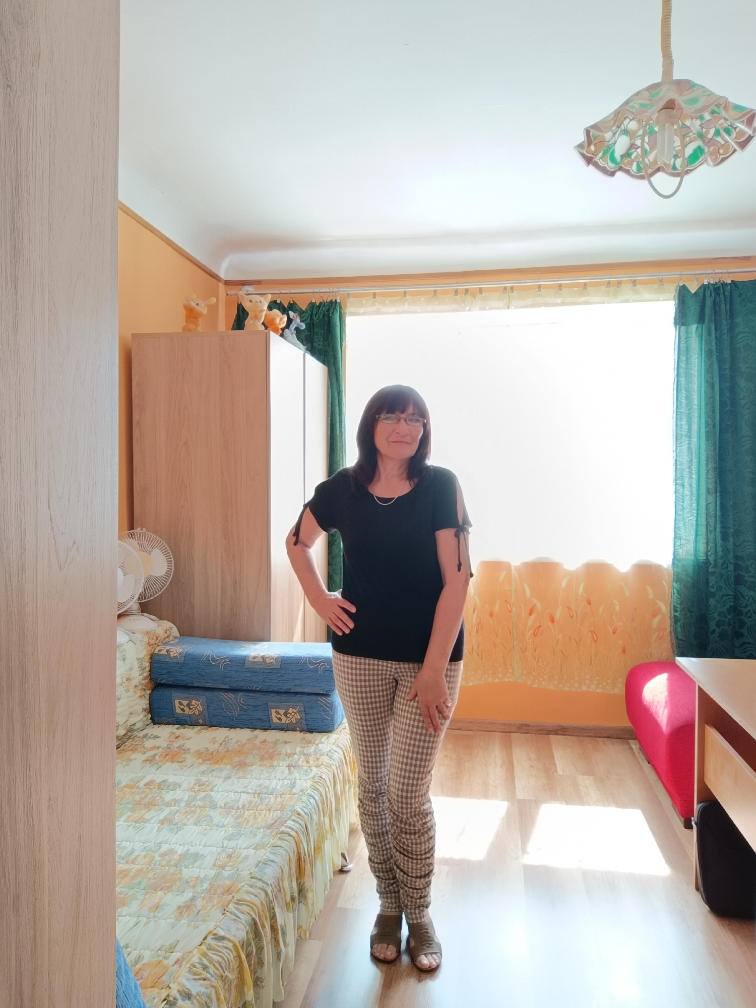 A woman stands in a room, facing the camera. She is wearing a black t-shirt and checkered pants. Her right hand is on her hip, and her left arm is relaxed by her side. The room appears to be a bedroom or living area. To the left of the woman is a bed covered with a floral patterned comforter and two blue pillows stacked on top. A wooden wardrobe is to the left of the bed. In the background, a large window is covered by sheer yellow curtains and dark green drapes. The floor is made of light-colored wood. A red sofa is visible to the right of the window. A desk is partially visible to the far right. The ceiling has a decorative light fixture. The overall impression is of a domestic interior. There is no indication that this image was taken in Tebras, Latvia.