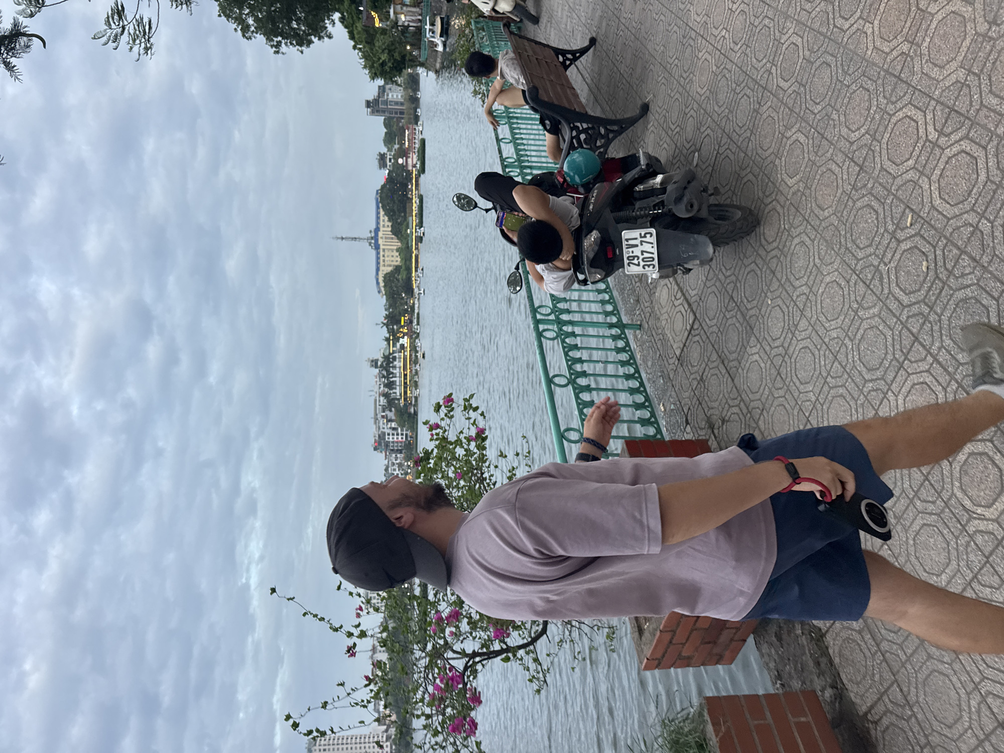 An overcast day in Hanoi, Vietnam, is depicted with a wide shot of West Lake. In the foreground, a man with a beard and a black baseball cap walks to the left, wearing a grey t-shirt and blue shorts, holding a black smartphone with a red strap in his right hand. Behind him, a green metal railing lines the paved walkway.
Further along the walkway, a motorcycle is parked. On the motorcycle, a person wearing a white t-shirt and black pants is resting with their head on their arms. To the right of the motorcycle, a wooden bench is occupied by two individuals. One person, wearing a grey t-shirt and dark shorts, is leaning back with their legs extended. Another person, partially visible and wearing a white shirt and dark shorts, is also seated.
The lake extends into the background, dotted with numerous small boats. The opposite shore is lined with buildings, including a tall tower with a pointed top. Trees and more buildings are visible along the distant coastline, under a cloudy sky. The walkway features a patterned tile surface.