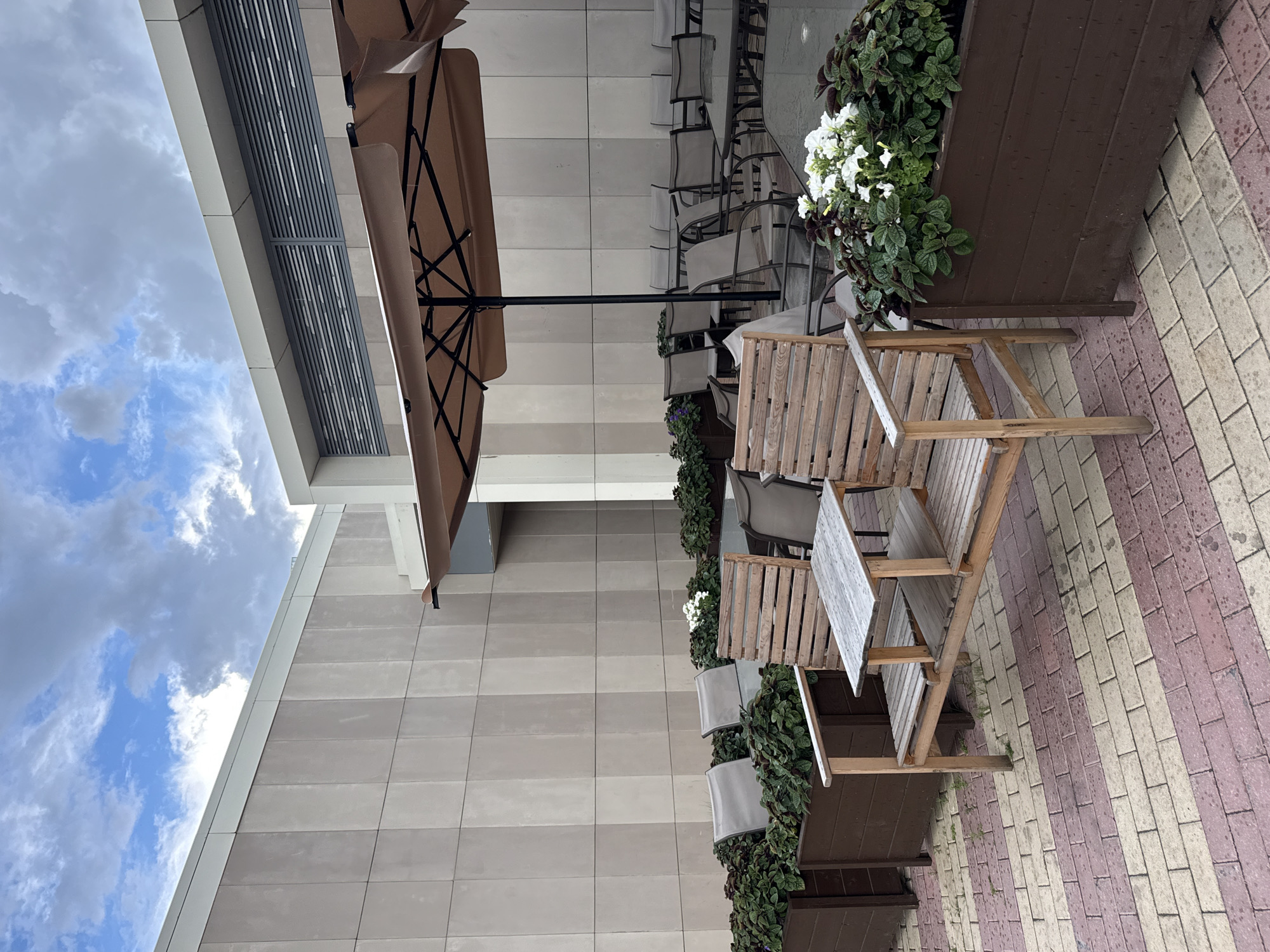 An outdoor seating area is shown under a cloudy sky, featuring a large tan umbrella providing shade over several tables and chairs. The area is paved with red and cream colored bricks. In the foreground, a wooden bench with a small attached table sits next to a dark brown planter filled with dark green foliage and white flowers. Behind this, a row of grey chairs and dark tables are visible, further back in the seating area. The background consists of a modern building facade with light beige square panels and horizontal grey vents. The scene appears to be a restaurant or cafe patio.