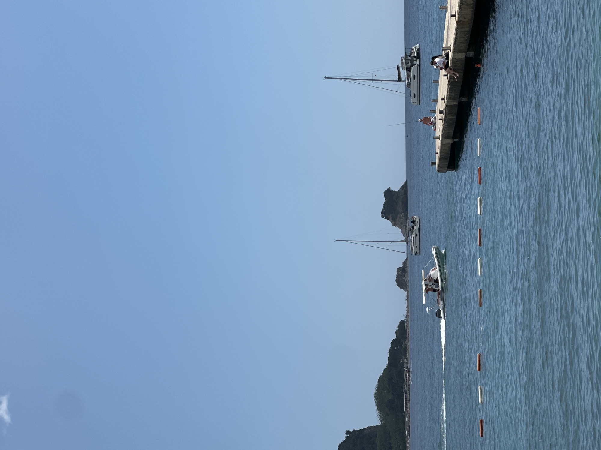 In the city of Budva, Montenegro, a bright, sunny day is depicted with a clear blue sky above. The scene is dominated by the azure Adriatic Sea. In the foreground, a line of orange and white buoys float on the water's surface. To the right, a concrete pier juts out into the sea, with two individuals seated on its edge. One person is wearing a white shirt and dark pants, while the other is in a black top and dark bottoms. Further along the pier, another individual is fishing.
The middle ground features several boats. A white speedboat with a canopy is cutting through the water, leaving a wake behind it, with three people visible on board. Two sailboats are anchored further out at sea. One is a monohull sailboat with a tall mast, and the other is a white catamaran. In the background, a lush, green, mountainous coastline with scattered buildings is visible. Two small rocky islets rise from the sea in the distance.