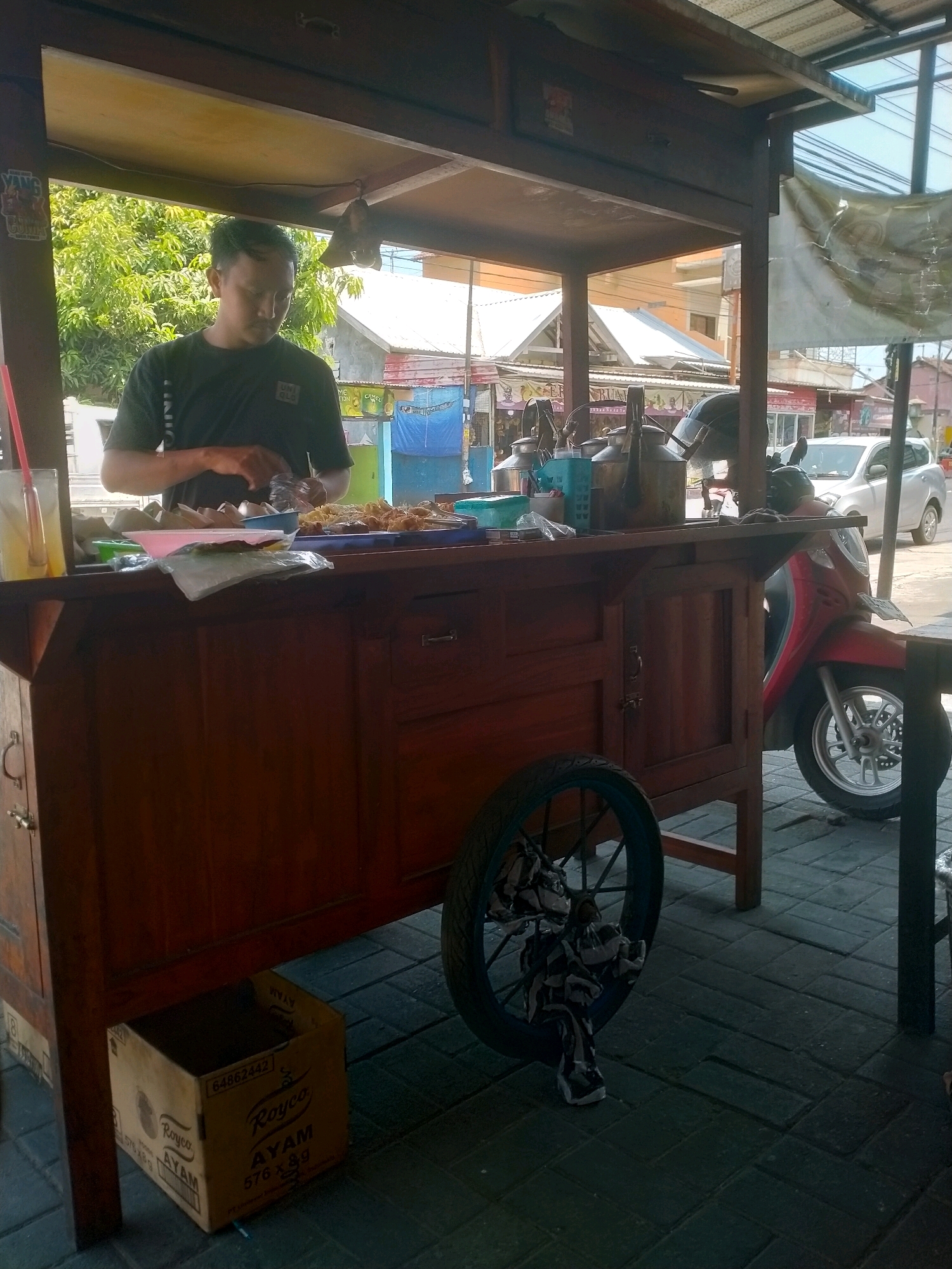 In Kasihan, Indonesia, a vendor stands behind a wooden food cart, preparing food. The vendor is a male wearing a black t-shirt and has short, dark hair. He is focused on the food on the cart, which appears to be fried items. The food cart is brown and has drawers and shelves. On the left side of the cart, there is a glass with a yellow drink and a red straw. In front of the cart, there is a red scooter. To the left of the cart, a cardboard box with "Royco AYAM" printed on it is visible. The scene is outdoors, with trees and buildings in the background, indicating a street-side setting. The ground is paved with dark tiles.