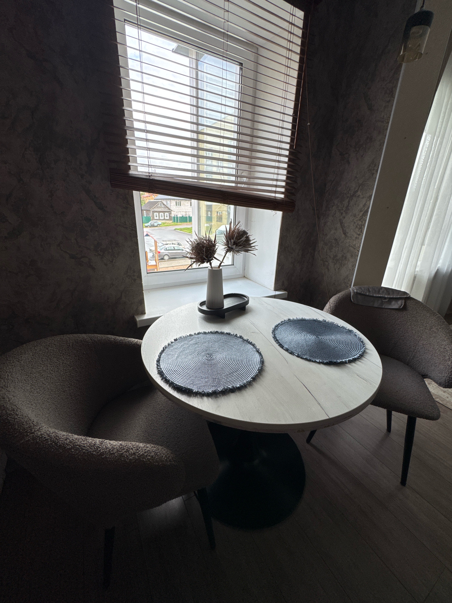 This is a medium shot, eye-level, indoor photograph of a dining set with two chairs and a circular table. The room has textured, dark grey walls. A large window with horizontal wooden blinds is positioned behind the table, revealing a view of houses, a street, and a car. The table is light wood with a black pedestal base and is adorned with two circular placemats and a vase with dried decorative plants. The chairs are upholstered in a fuzzy, brown fabric and have dark legs. A white curtain is visible to the right of the window. The floor is made of dark wood planks. This scene appears to be in a residential setting in Valday, Russia. There are no people visible in the image.