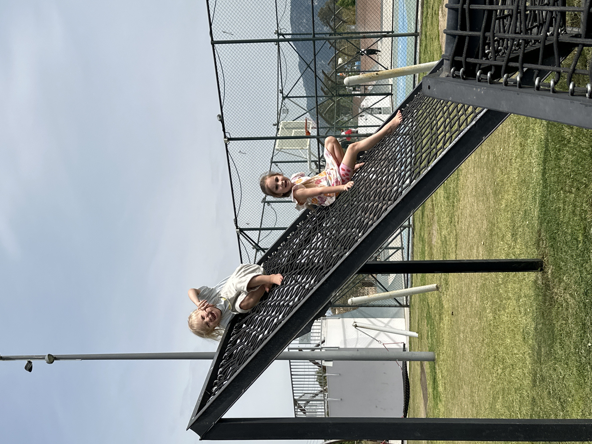 Two young girls are on a playground structure in Fethiye, Turkey. The structure appears to be a climbing frame with a net. One girl, dressed in a floral sundress, is lying on her back. The other girl, wearing a light-colored shirt, is further down the structure, and also lying on her back. The playground is located outdoors with a grassy area. In the background, a basketball court and netting surround the area. Mountains are visible in the distance, along with other buildings. The sky is overcast.