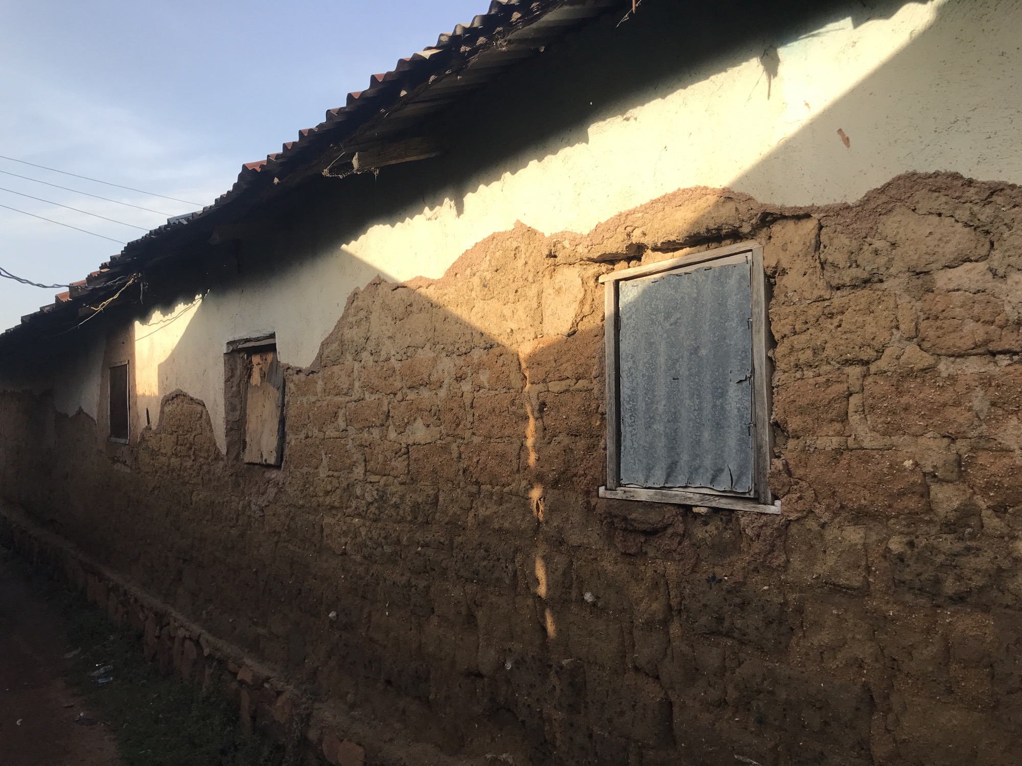 The image shows a close-up view of the exterior wall of a mud-brick building in Kabong, Nigeria. The wall is constructed from rough, reddish-brown mud bricks and appears weathered and uneven. Sunlight casts dramatic shadows across the wall, highlighting its textured surface. A corrugated metal sheet is visible in a window opening, and another smaller window to the left is boarded up. The roofline of the building, with its corrugated metal sheeting and exposed wooden beams, is visible at the top of the frame. The overall impression is of a humble, rural dwelling. The surrounding environment is suggested by a dirt path leading away from the wall and some sparse vegetation.