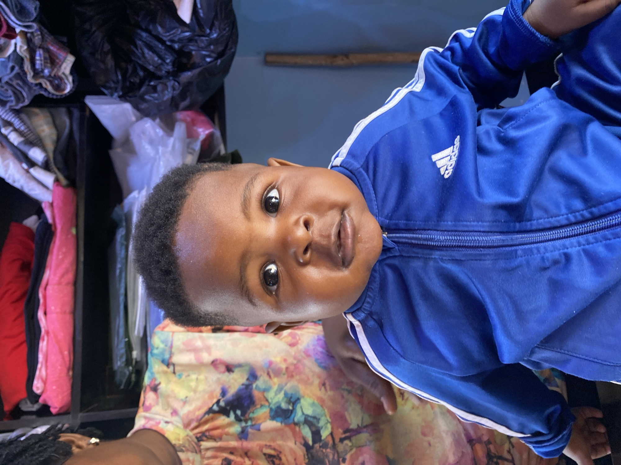 A young child with dark skin and short hair is wearing a blue Adidas jacket with white stripes down the sleeves. The child is smiling slightly and looking directly at the camera. The background shows a cluttered indoor space. To the left is a dark, full closet containing items of clothing wrapped in plastic and stuffed inside. A partially visible adult is wearing a colorful floral top. The location is Bukuru, Nigeria.