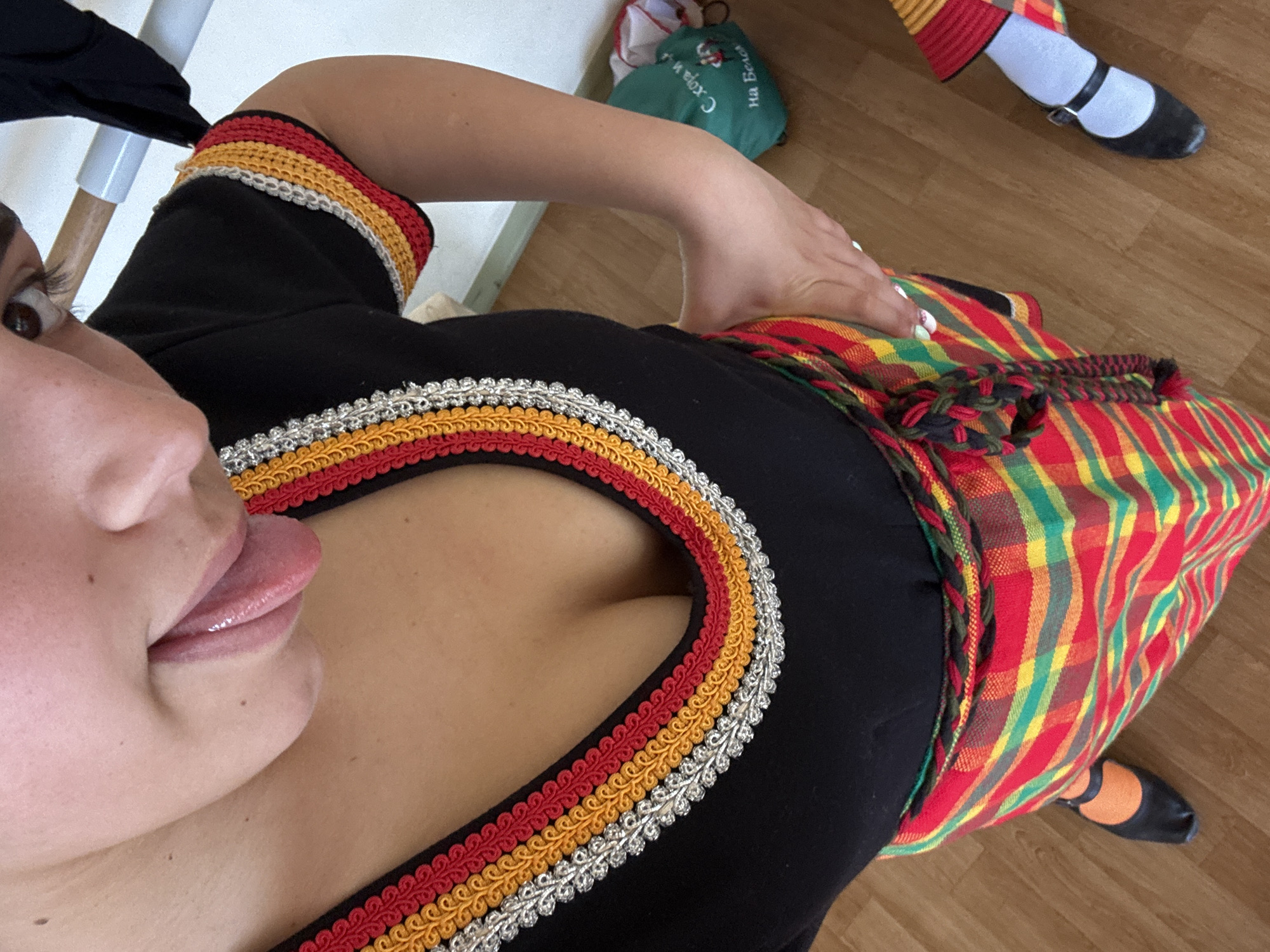 A close-up, high-angle shot captures a woman wearing a black shirt with red, yellow, and silver trim around the neckline, along with a plaid skirt in red, yellow, and green. The woman's face is partially visible, with her tongue sticking out. Her hand rests on the skirt. Another person is partially visible in the upper right corner, wearing striped socks and black shoes. A green bag with lettering is on the floor. The scene is indoors, set in an unknown location.
