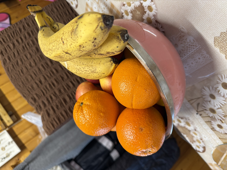 The image displays an indoor still life, reportedly located in Tarnava, Bulgaria. A pink bowl with a metallic rim, containing assorted fruits, is positioned on a light-colored tablecloth adorned with a pattern of white daisy-like flowers and lace trim. The prominent fruits include three distinct oranges, notable for their textured, bright orange peels, situated towards the left and center of the bowl. A bunch of yellow bananas, exhibiting brown speckles indicative of ripeness, extends from the right side of the bowl. Beneath and behind these, several red apples and other fruits are partially visible within the bowl. The background is blurred, revealing light brown wooden wall paneling to the upper left, a folded grey garment, and a dark brown textured fabric item to the upper right. No individuals or explicit actions are visible within the frame.