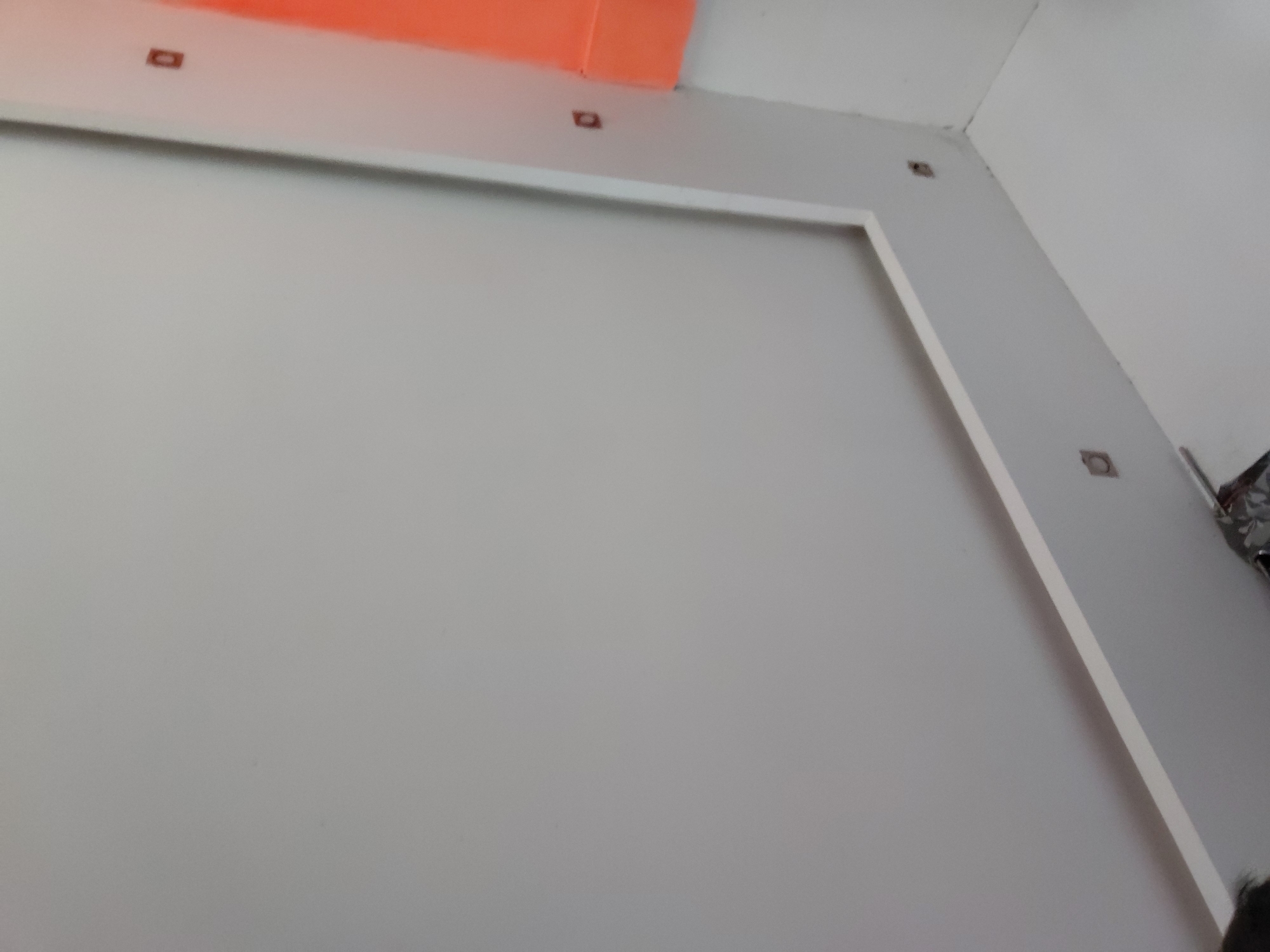 The image displays a bright, clean interior space, likely a room under construction or recently renovated, located in Santa Ana, Venezuela. The dominant feature is a large, light-grey wall, which is partially bordered by a thick, white, rectangular trim or paneling on its top and right sides, creating a defined inset or framed area.
Above the main wall, a section of a vibrant orange ceiling or beam is visible on the left, adding a distinct color contrast to the otherwise neutral palette of white and light grey. This orange section meets a white ceiling. Small, dark, square or rectangular cutouts are noticeable on the white surfaces, specifically along the top edge of the framed wall and on the white ceiling segment near the orange area. Another similar cutout is seen on the right wall, embedded within the white trim. These cutouts appear to be unfinished openings, potentially for light fixtures, outlets, or other installations.
Towards the upper right, the corner where two white walls meet is visible. Along the far right edge of the frame, some indistinct metallic or reflective objects are partially visible, and a small segment of dark hair can be seen at the bottom right corner, suggesting the presence of a person just outside the frame.
There are no people prominently featured, nor any specific activity taking place. The scene is static, highlighting architectural details. The lighting suggests an indoor setting, without clear indicators of the time of day or outdoor weather. No visible text is present in the image.