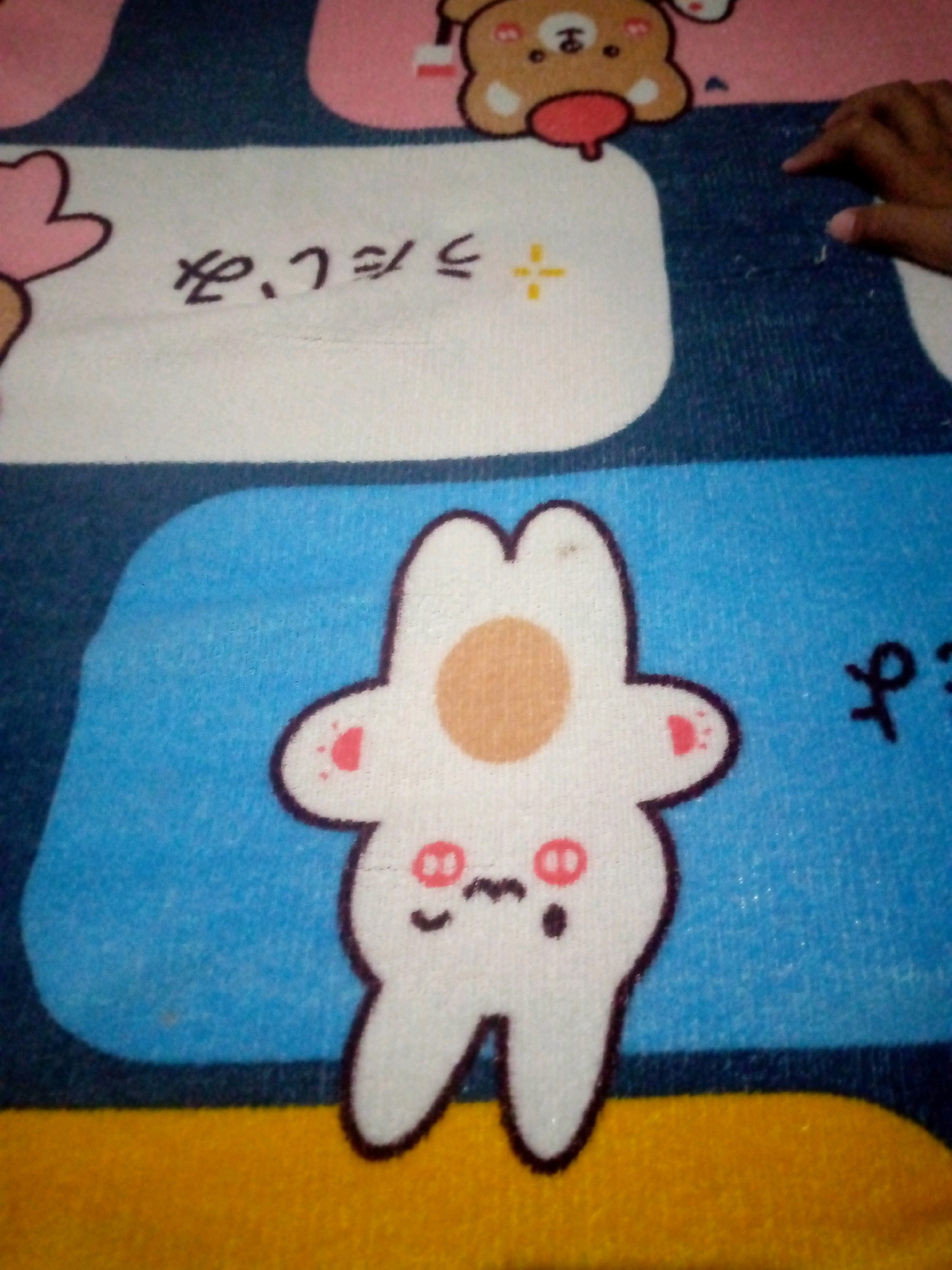 An overhead medium shot captures a close-up view of a fuzzy, white, cartoon-like rabbit figure with a brown circle on its head, pink blush marks on its cheeks, and a simple facial expression. The rabbit is situated on a blue surface, which is part of a larger patterned fabric. In the background, a white rectangular area contains dark Japanese writing. To the right of the rabbit, a hand with visible fingernails is partially visible, with fingers extended towards the fabric. The overall impression is that of a playful or decorative item. The image's context in Sumberwono, Indonesia is not discernible from the visual information provided.