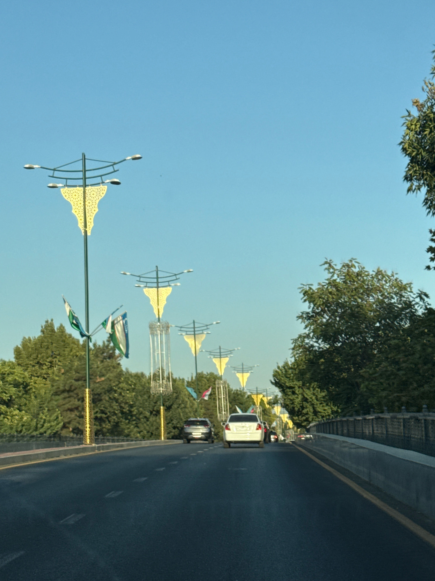 The image depicts a street scene in Tashkent, Uzbekistan, with multiple ornate streetlights and flags lining the road. The streetlights feature yellow decorative elements and are spaced at regular intervals. Several flags, appearing to be the national flag of Uzbekistan with its distinct blue, white, and green stripes and crescent moon and stars, are visible on poles near the streetlights.
The road is paved with asphalt and has white dashed lane markings. A few cars are driving on the road. In the foreground, the road surface is dark. In the middle ground, there are trees and a bridge or overpass with a railing. The sky is clear and blue, suggesting a sunny day. The overall impression is one of a well-maintained urban environment. The specific location within Tashkent is not identifiable from the visual cues alone, but the presence of national flags and distinctive streetlights suggests a public or ceremonial area.