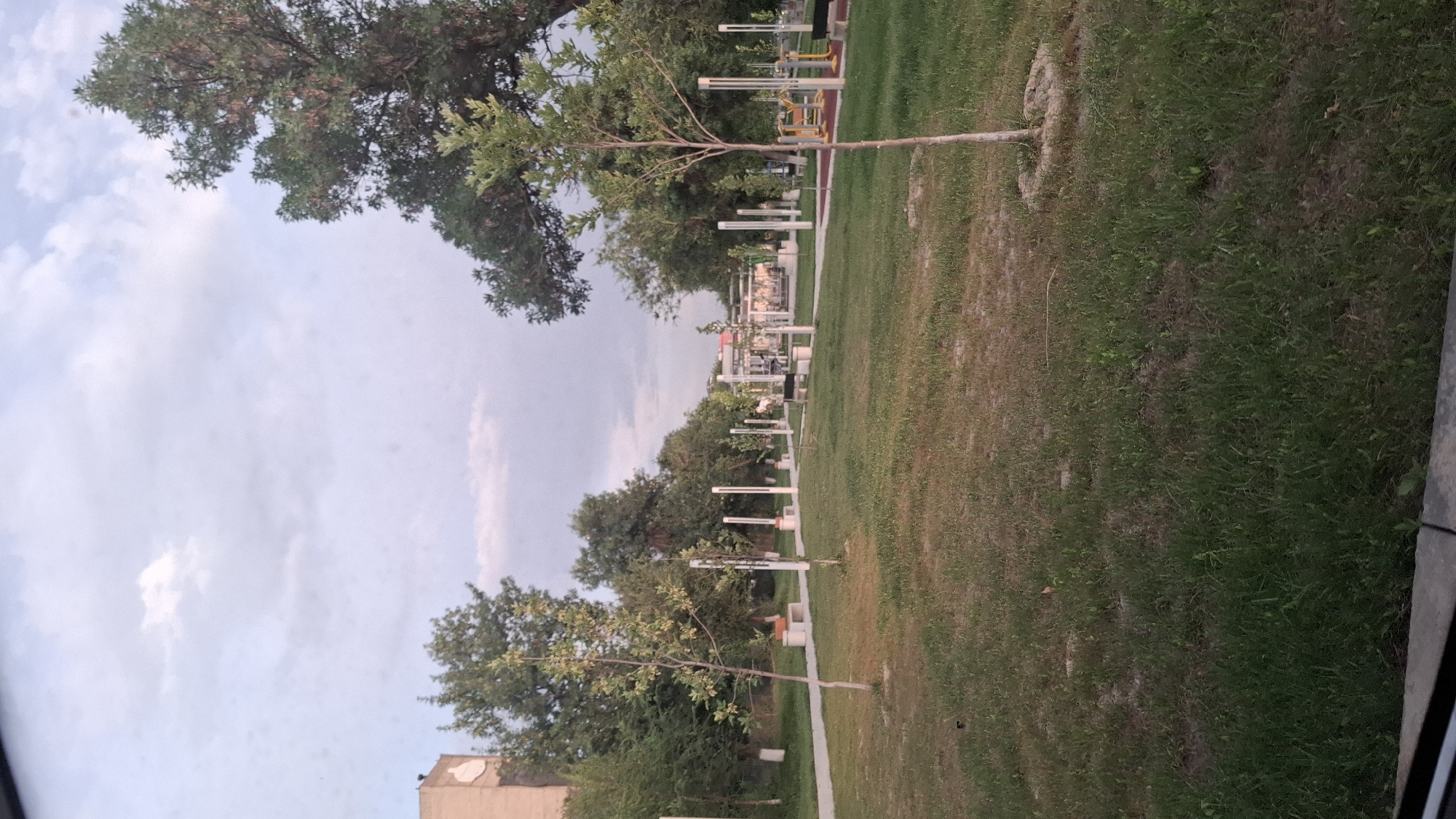 This is an outdoor photograph of a park area in Shymkent, Kazakhstan. The scene is dominated by a large grassy lawn with scattered trees. In the foreground, a young tree stands to the right, and a paved pathway curves through the grass to the left. Further back, a line of white poles with lights is visible, alongside park benches and a few pieces of outdoor exercise equipment, including yellow-framed machines. The background features more trees and a building on the far left. The sky is overcast with scattered clouds. The overall impression is of a public recreational space.