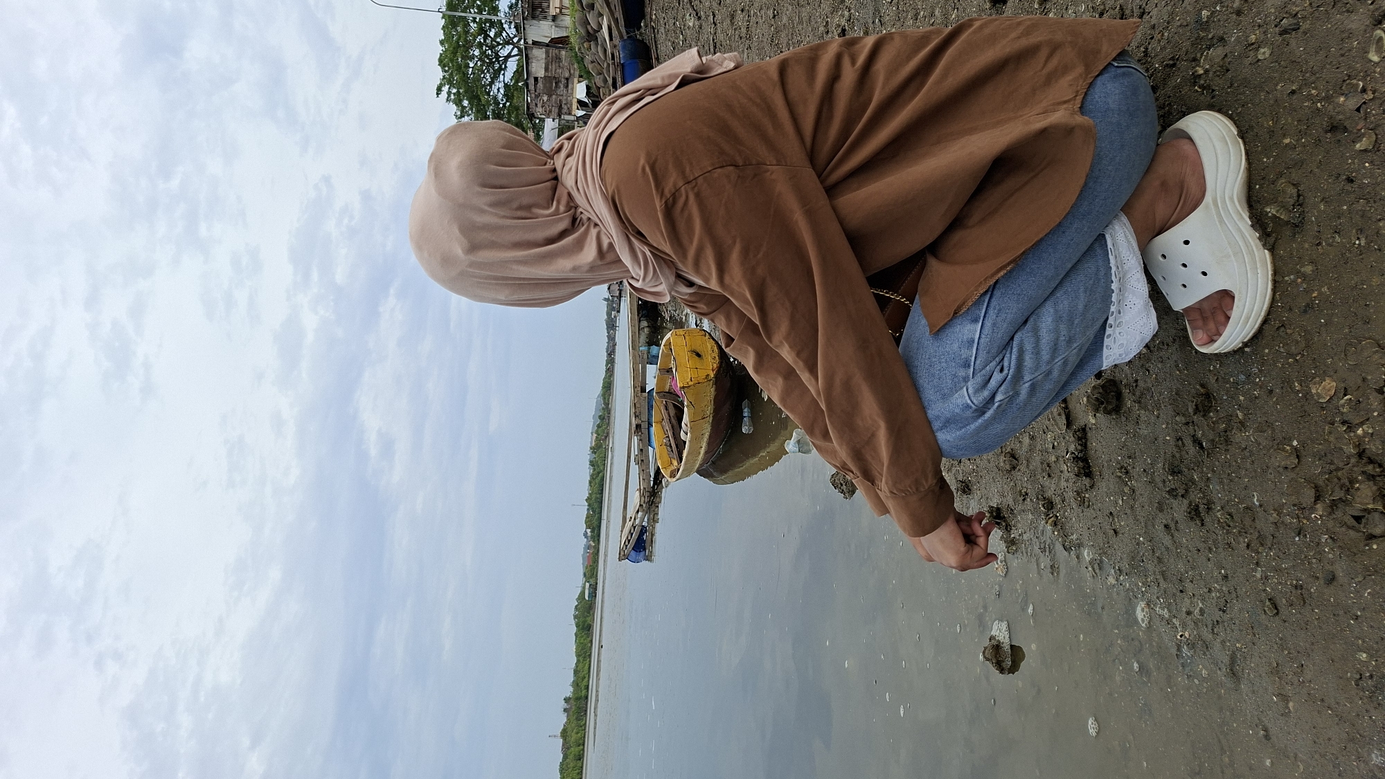 A person wearing a light brown hijab and a brown long-sleeved shirt is crouching on a muddy shoreline, facing away from the camera and looking out at a calm body of water. The person is wearing blue jeans with white lace trim visible at the ankle and white platform slides. In the water, there is a small yellow and brown boat tied to a wooden dock. The background shows a line of trees and some buildings along the distant shore, suggesting a coastal or riverine environment. The sky is overcast with light gray clouds. This scene is likely in the city of Lhokseumawe, Indonesia, based on the presence of such coastal landscapes and vernacular architecture.