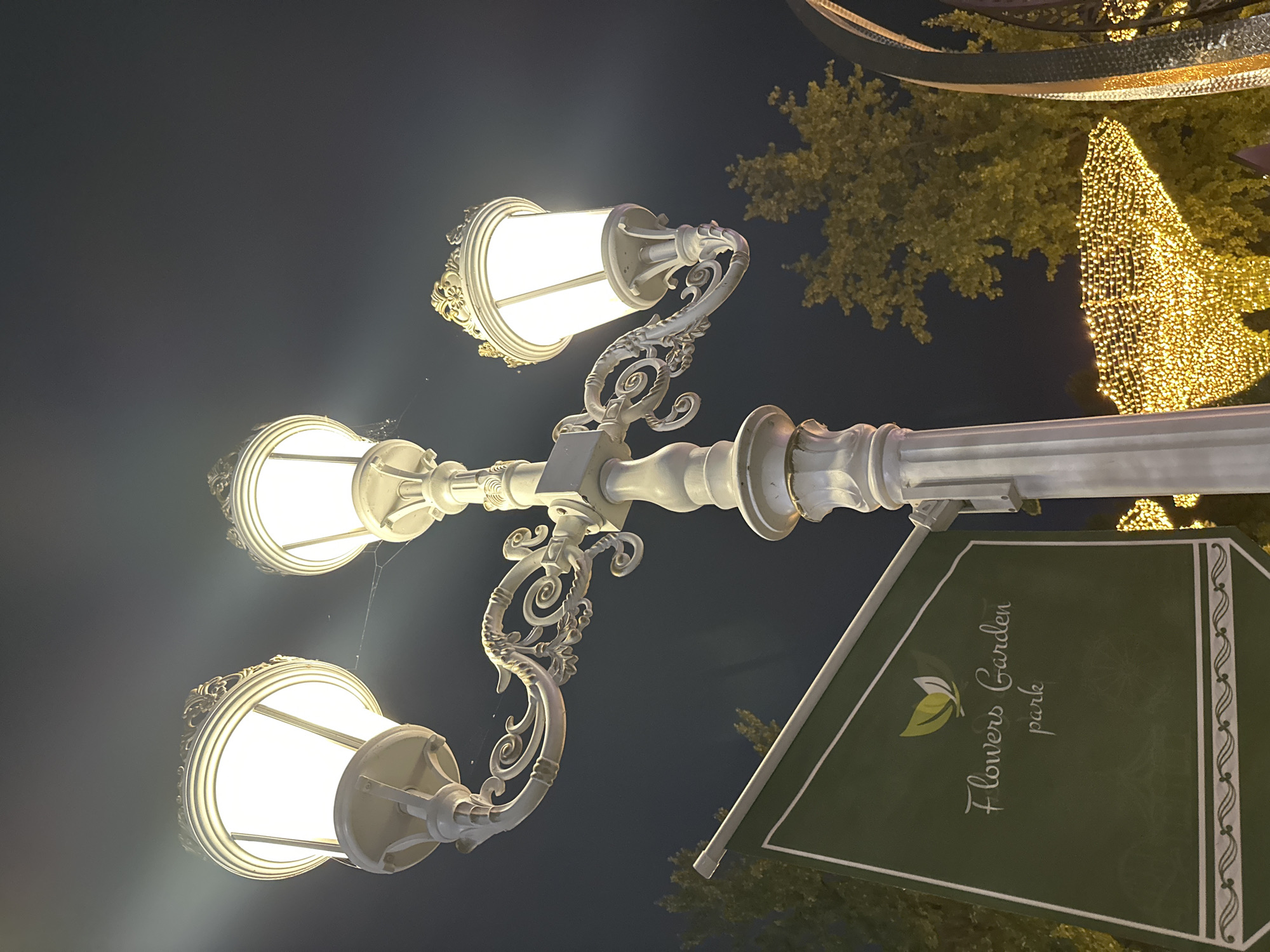 An ornate, white, triple-headed lamppost stands against a dark night sky. The lamppost has decorative scrollwork and ornate finials on each lamp housing. One lamp is positioned to the left, another in the center, and a third to the right, all emitting a bright, warm light. A delicate spiderweb is visible between the two leftmost lamps.
To the left of the lamppost, a dark green banner is displayed, featuring a light green logo of two leaves. Below the logo, in white script, it reads "Flowers Garden park".
In the background, trees with yellowing leaves are illuminated by soft lights, and a cluster of warm yellow string lights is visible, suggesting a festive or park-like setting. Part of a decorative metal structure is visible on the far right. The overall scene depicts an illuminated park or garden area at night. The sign indicates this location is likely "Flowers Garden park" in Tashkent, Uzbekistan, given the provided context.