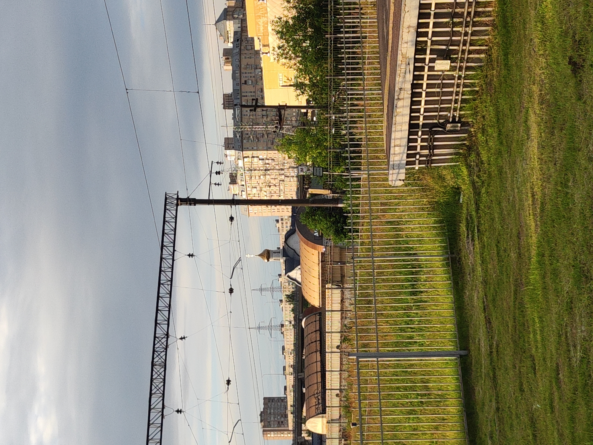 The image depicts an urban railway landscape under a cloudy sky. In the foreground, a green grassy area is bordered by a metal fence. Beyond the fence, a railway platform with a textured surface is visible. The middle ground features several industrial-style buildings with curved roofs, possibly railway depots or maintenance sheds. A church with a golden dome and cross stands out among these structures. In the background, a series of multi-story apartment buildings of varying architectural styles form the urban skyline. Overhead, railway catenary wires and supporting structures crisscross the sky, indicating an active rail line. The lighting suggests it is either early morning or late afternoon, with the sun casting warm light on some of the buildings. The scene is devoid of people. This appears to be a view of a railway station or yard in Saint Petersburg, Russia, possibly near the city's residential areas.