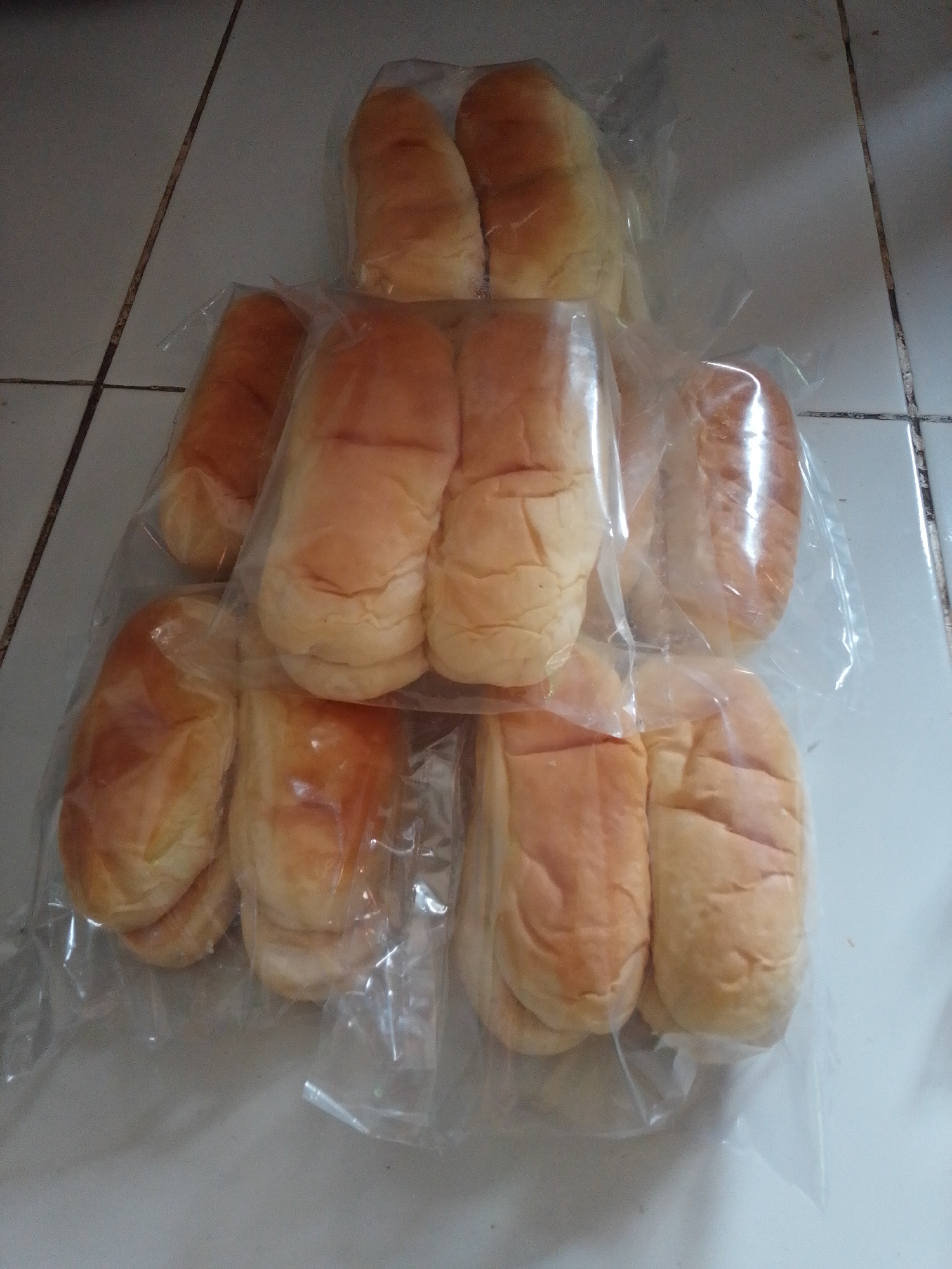 A group of individually wrapped, golden-brown bread rolls are arranged on a tiled surface. The bread rolls are soft and appear to be a sweet variety. The tiles are light-colored with dark grout lines, suggesting an indoor setting, possibly a kitchen or food preparation area. The lighting is dim, creating soft shadows. The image is taken from a slightly elevated perspective.