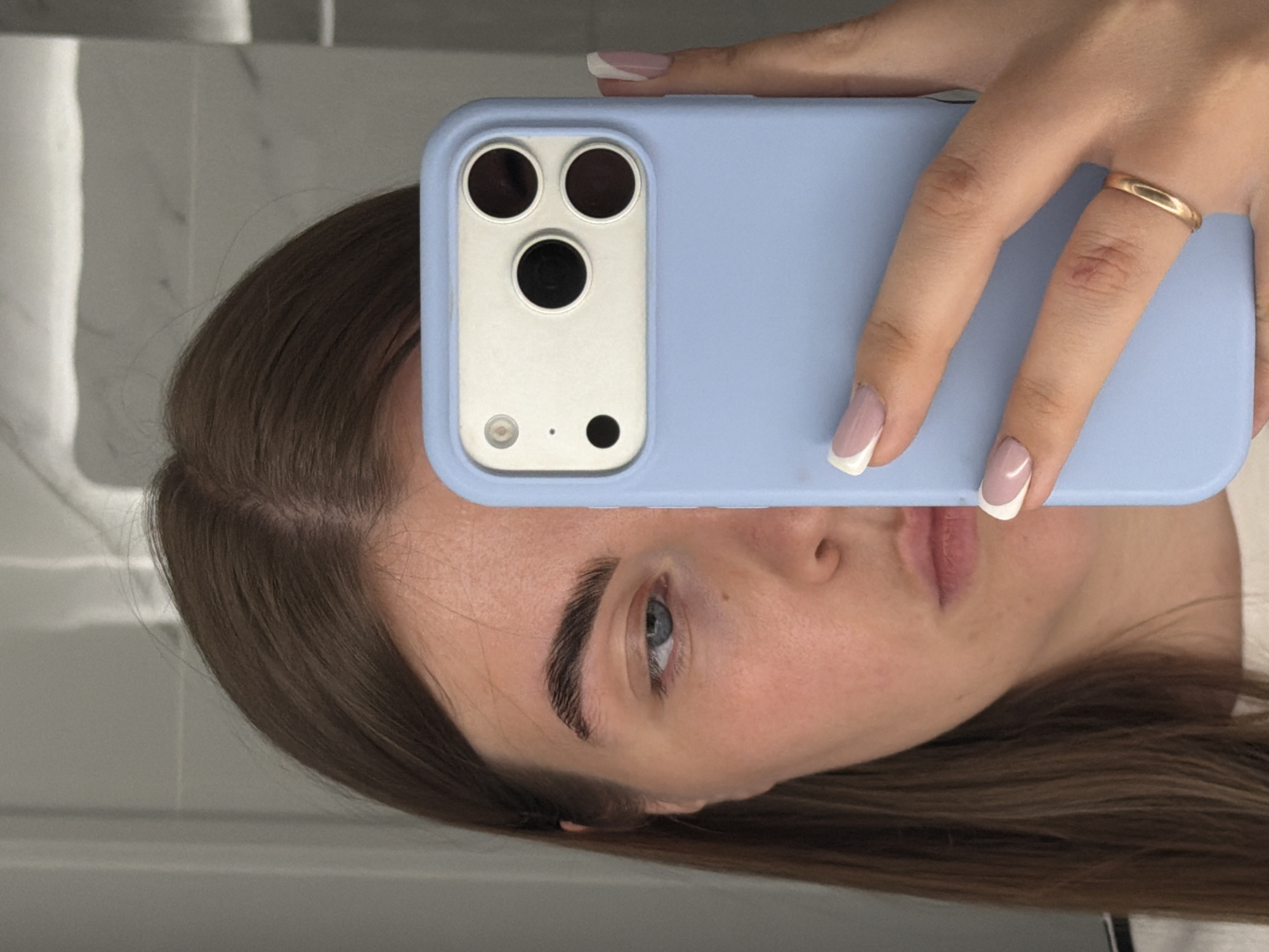 A woman holds a blue phone case with three camera lenses in front of her face. Her left eye and part of her eyebrow are visible above the phone. She has fair skin, brown hair, and wears a gold ring on her finger. She has white-tipped French manicured nails. The background is a light gray wall, possibly a bathroom. There is no indication of the city Chaikovsky, Russia in the photo.