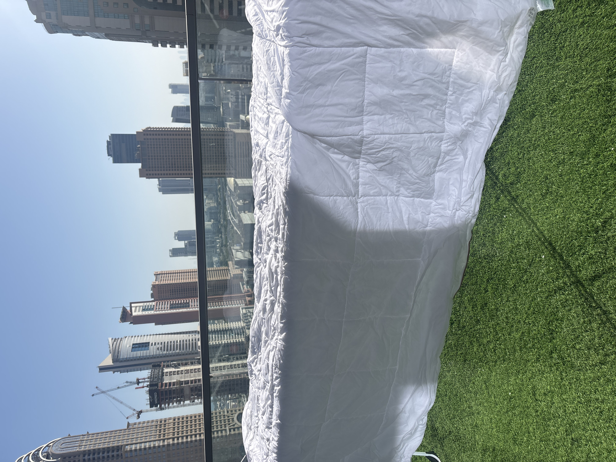 A white duvet is laid out on a balcony with a green artificial turf floor. In the background, a cityscape with numerous skyscrapers and construction cranes is visible under a clear blue sky. The buildings appear to be in Dubai, United Arab Emirates, characterized by modern architectural designs and high-rise structures. The duvet is draped over a structure that is not fully visible, possibly a clothes drying rack or a table. The sunlight creates shadows on the duvet, indicating it is daytime. A glass railing separates the balcony from the view of the city.