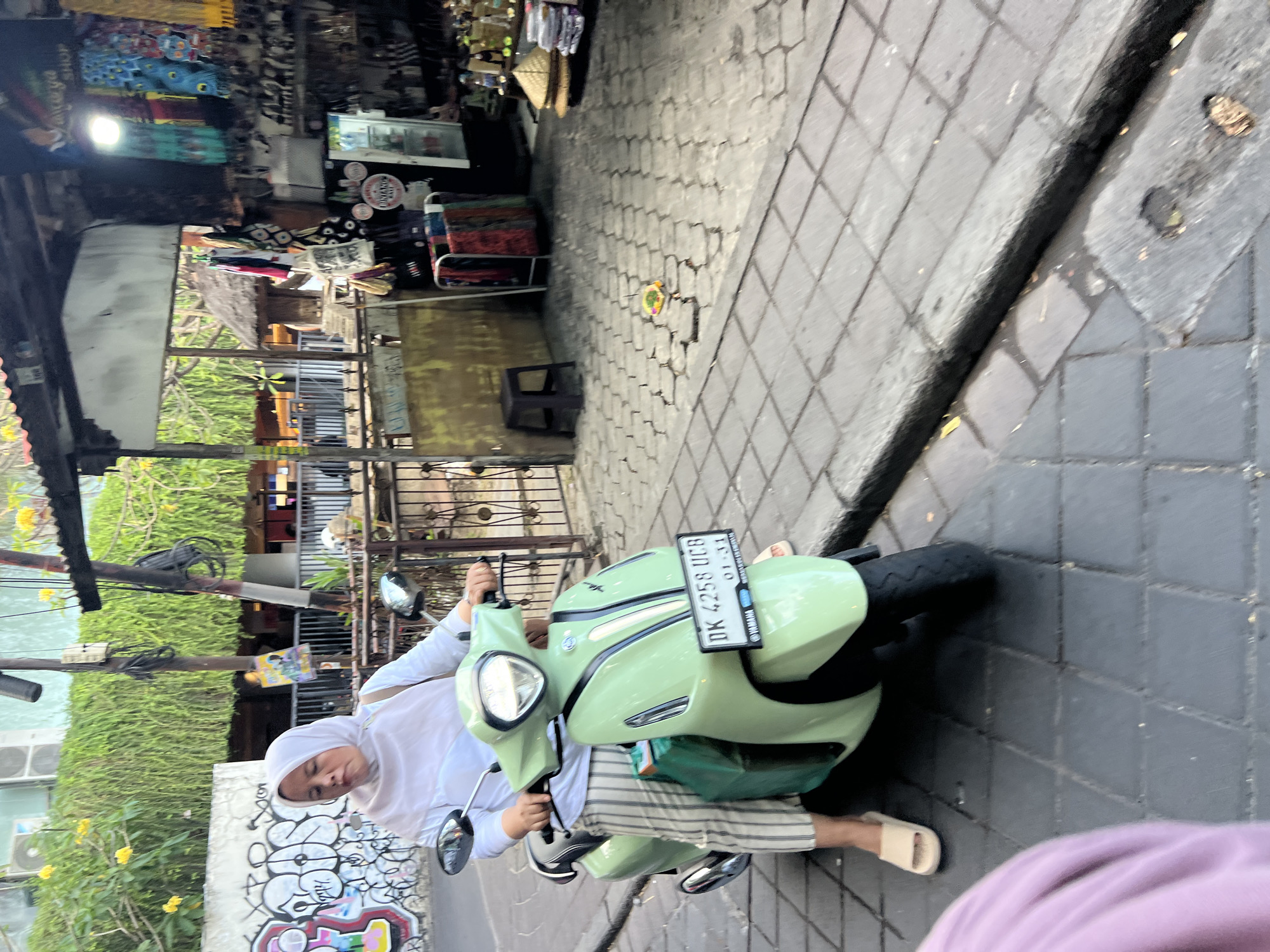 A woman wearing a white hijab and a white long-sleeved top, along with vertically striped pants, sits on a light green scooter with a license plate reading "DK 4258 UCB 01-31". She is on a paved street in Kuta, Indonesia. The street is lined with shops and buildings. In the background, a shop displays colorful fabrics and souvenirs. Lush green vegetation hangs from a building on the left. The ground is paved with hexagonal and square tiles, with a small colorful offering on the ground.