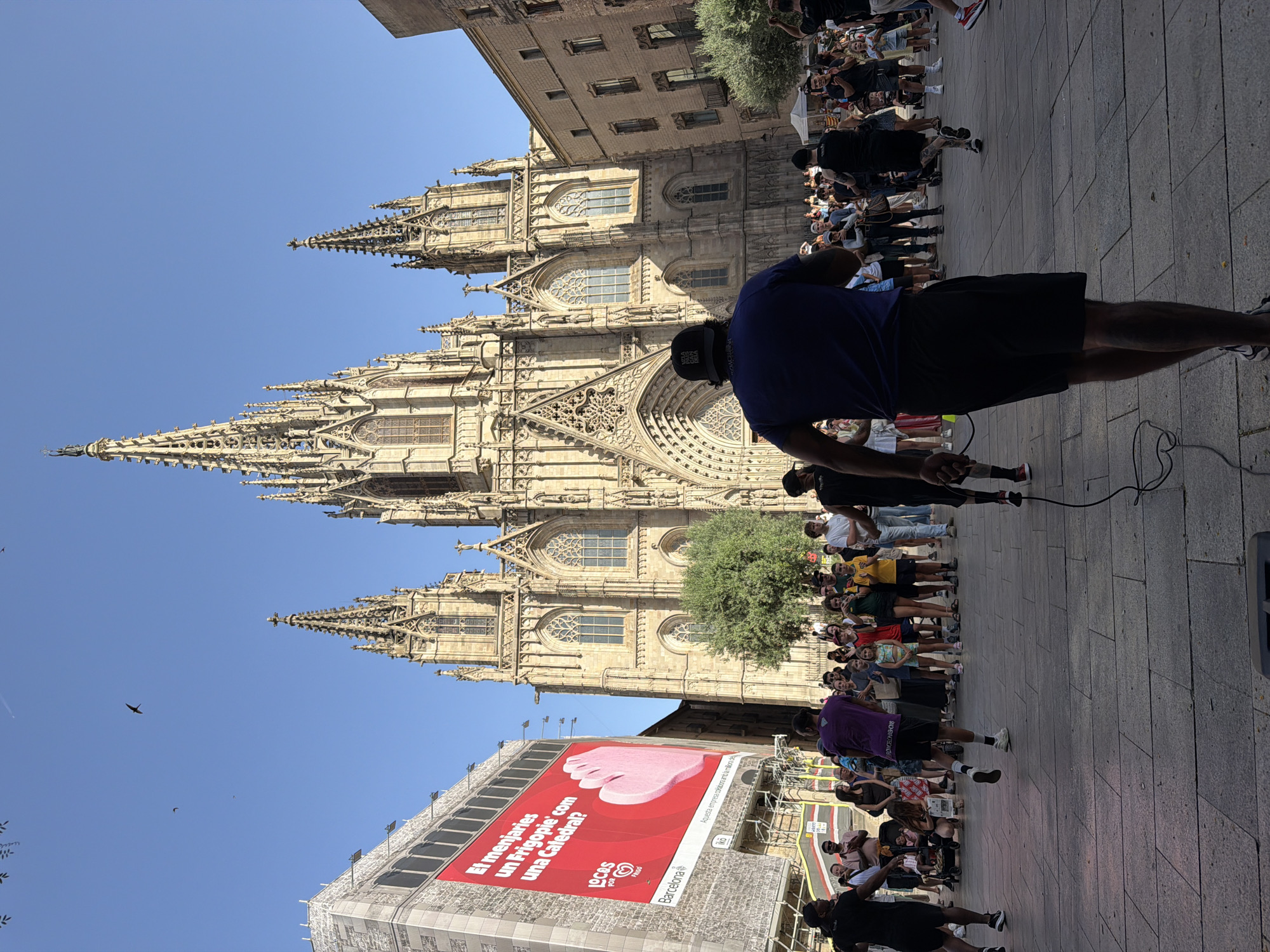 The image captures a vibrant scene in front of the Barcelona Cathedral in Spain. In the foreground, a group of people are gathered, some appearing to be participating in an outdoor fitness activity, possibly jump roping, as evidenced by the visible ropes and athletic attire. The main subject in the foreground is a man wearing a blue t-shirt and black shorts, facing away from the camera, holding a jump rope. Behind him, a crowd of spectators and participants look towards the majestic Gothic architecture of the Barcelona Cathedral, a prominent landmark. The cathedral's spires and intricate facade are illuminated by the sun under a clear blue sky. To the left, a large billboard with a red background displays an advertisement in Catalan, featuring a hand-shaped graphic and text that reads "Et menjaríes un Frigopie' com una Catedrál?" which translates to "Would you eat a Frigopie' like a Cathedral?". The overall atmosphere appears to be lively and social, with people enjoying a sunny day in a historic urban setting.