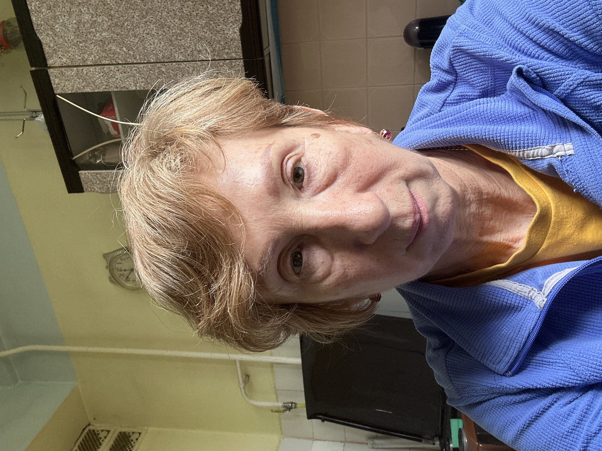 An older woman is taking a selfie indoors. She has short blonde hair and is wearing a blue jacket over a yellow top. The background includes kitchen cabinets, tiled walls, and a clock. The location cannot be determined as Moscow, Russia based on visual information alone.