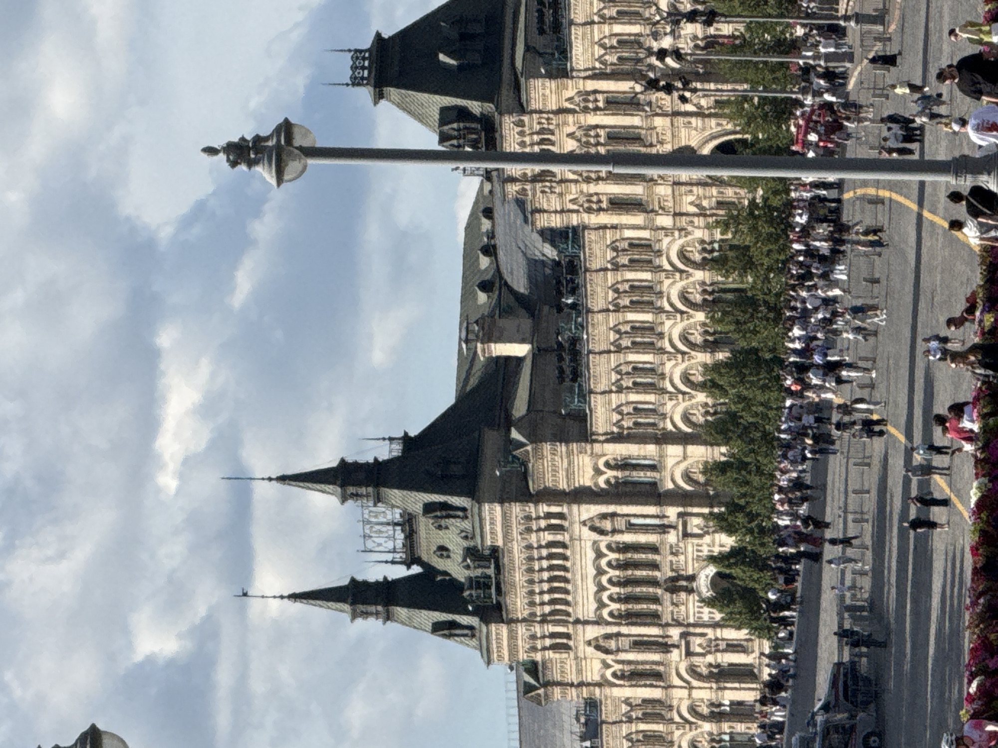 The image depicts a wide shot of Red Square in Moscow, Russia, under a cloudy sky. The iconic GUM department store, a large historical building with ornate architecture and distinctive green mansard roofs, dominates the background. Two prominent spires rise from the GUM building, adding to its grandeur. In the foreground, a busy public space is filled with numerous people walking and standing. Several lamp posts, including a tall, ornate one on the right side of the frame, are visible. A streetcar or bus is seen on the left, and various barriers or bollards are placed throughout the square. Trees line the area in front of GUM. The overall scene suggests a lively day with many visitors exploring the historic square.