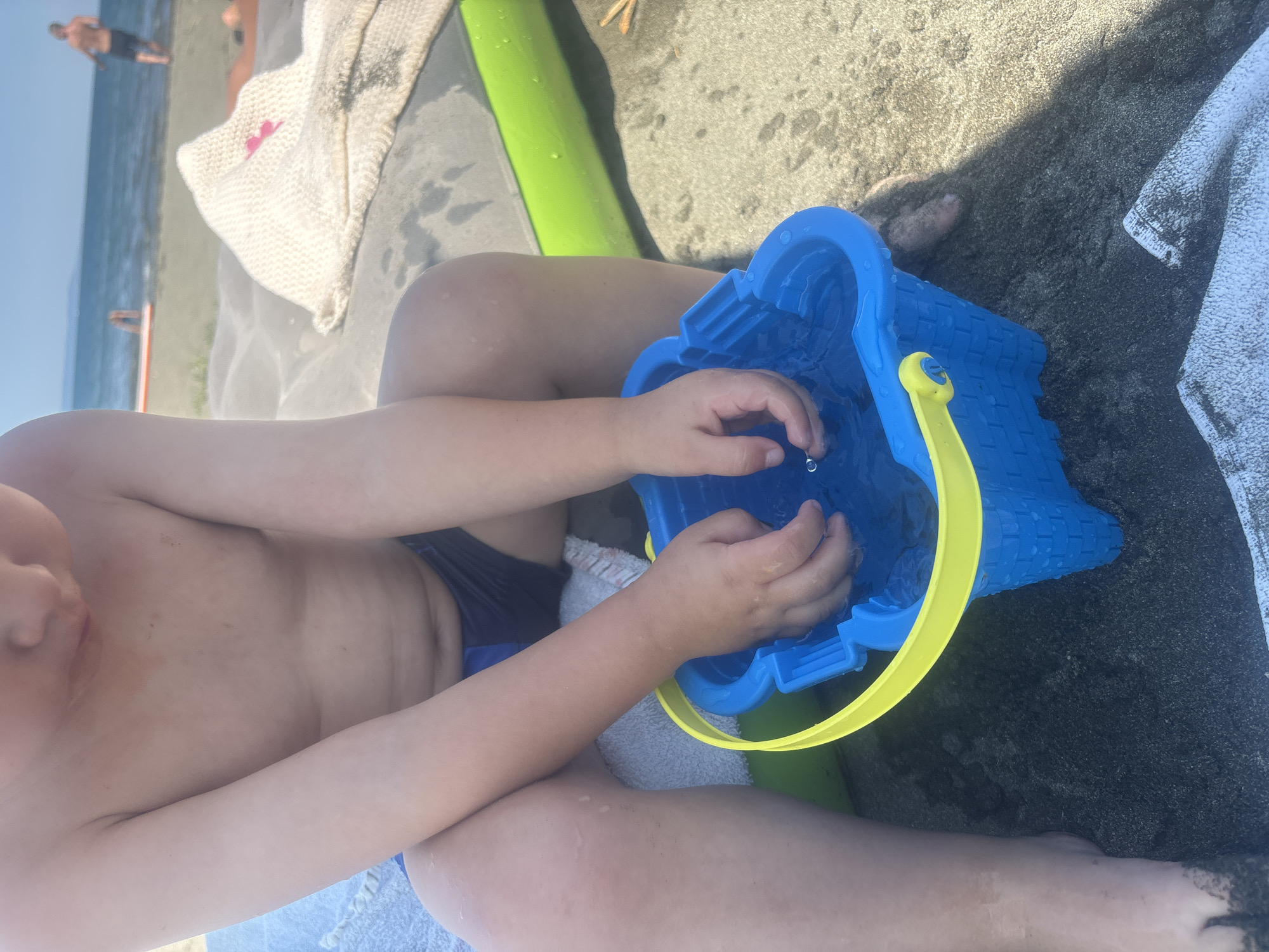 The image shows a close-up view of a child's hands interacting with a blue toy bucket filled with water. The child is wearing dark swim trunks and is sitting on dark sand. A bright green object, possibly a pool noodle, is partially visible behind the child. In the background, the sea and a beach are visible, with a few people in the distance. The lighting suggests it is daytime, likely sunny, given the beach setting. The overall scene depicts a child playing at the beach. There is no visible text in the image.